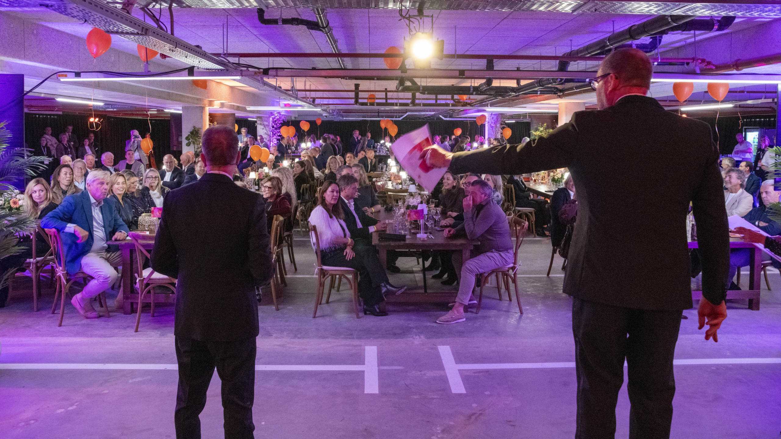 Een drukke zaal met mensen die aan tafels zitten onder kleurrijke verlichting, terwijl twee mannen het publiek toespreken vanaf de voorgrond.
