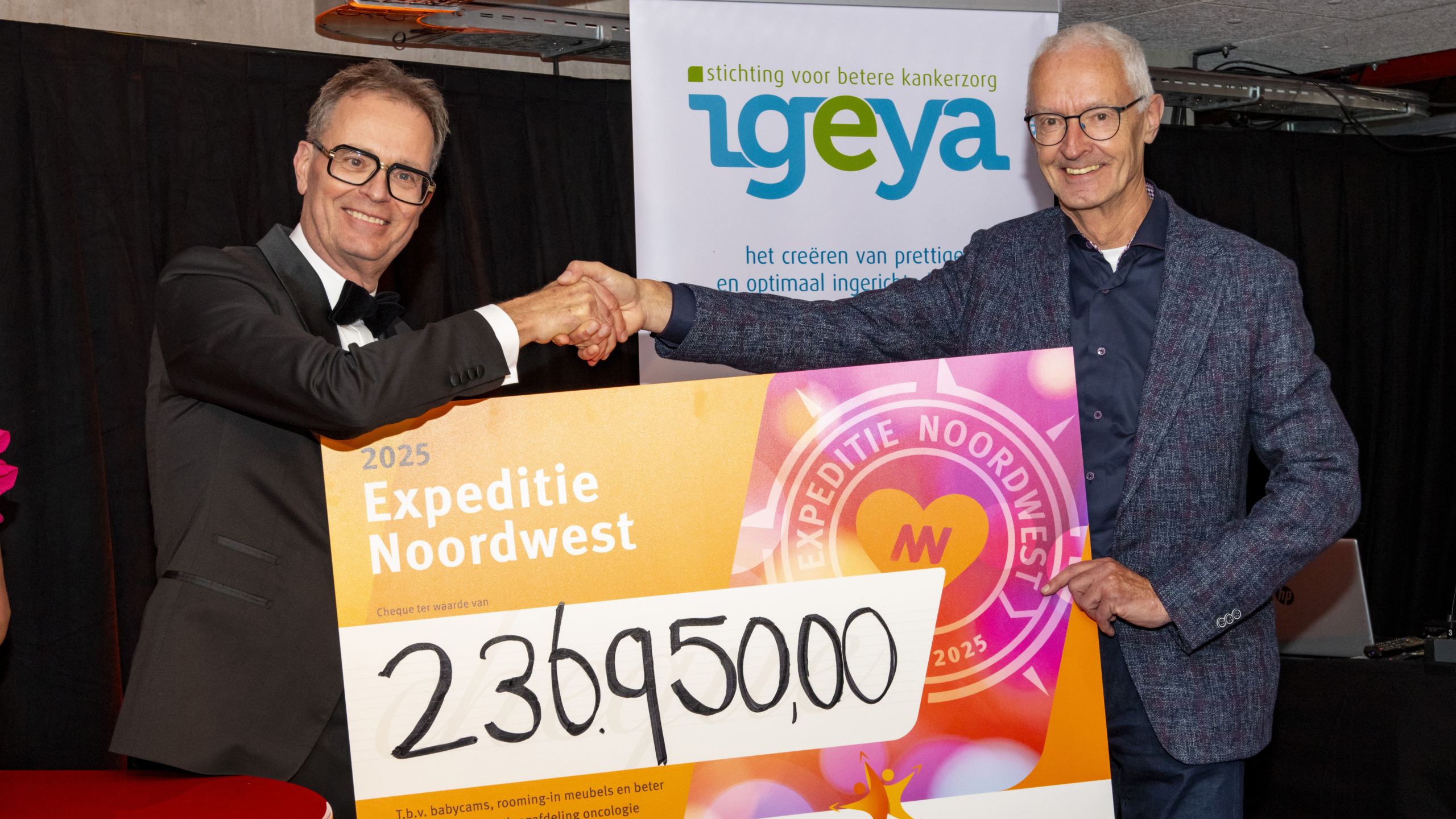 Twee mannen schudden handen terwijl ze een grote cheque vasthouden met het bedrag van 236.950,00 euro voor Expeditie Noordwest, in aanwezigheid van een bord met het logo van Stichting Igeya.