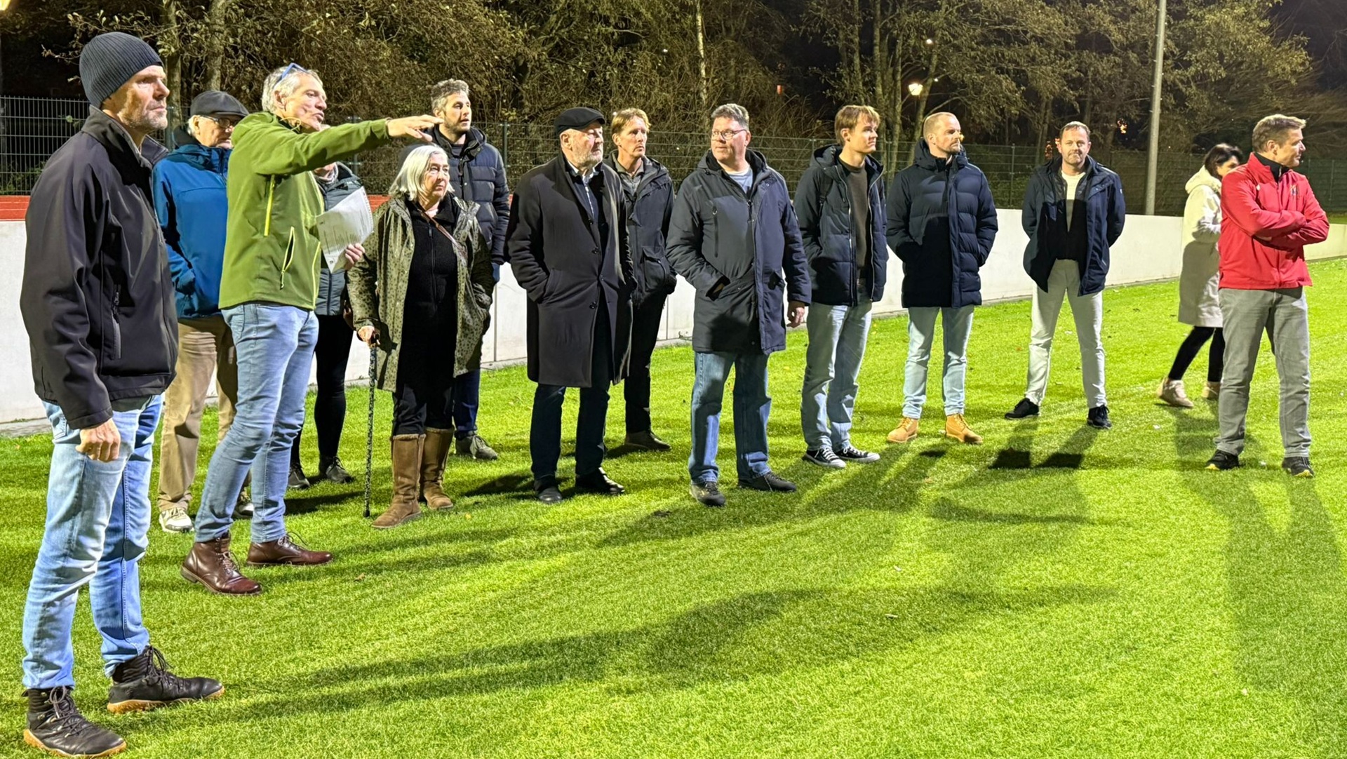 Groep mensen staat buiten op een grasveld, gekleed in warme jassen en laarzen, met één persoon die wijst naar iets in de verte.