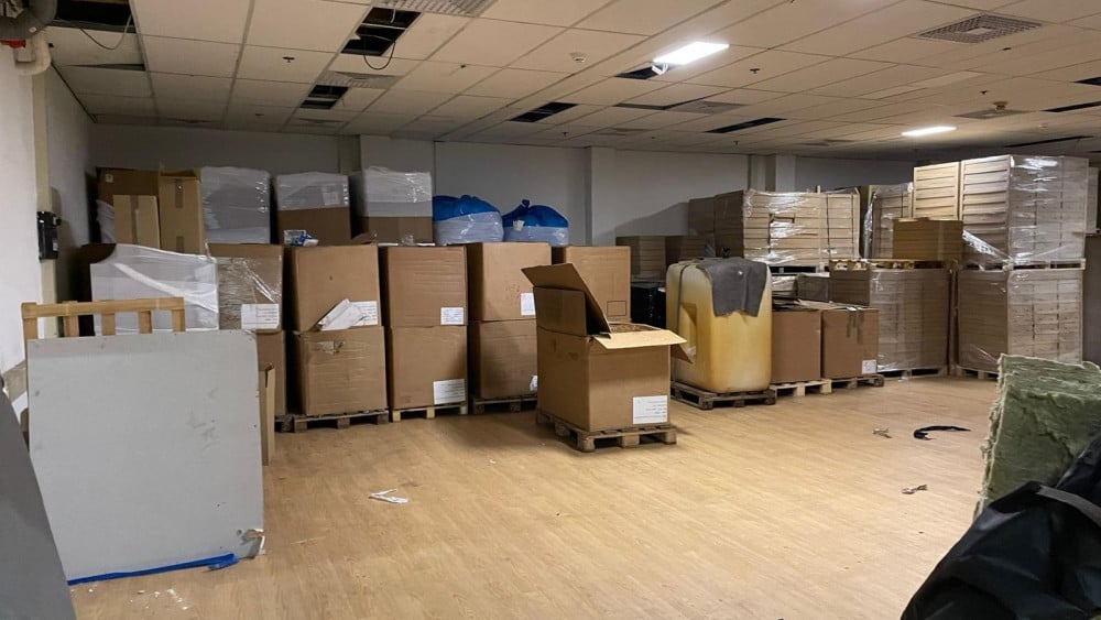 Opslagruimte gevuld met grote kartonnen dozen op pallets, sommige dozen zijn geopend. De ruimte heeft een systeemplafond met enkele ontbrekende platen en een houten vloer.