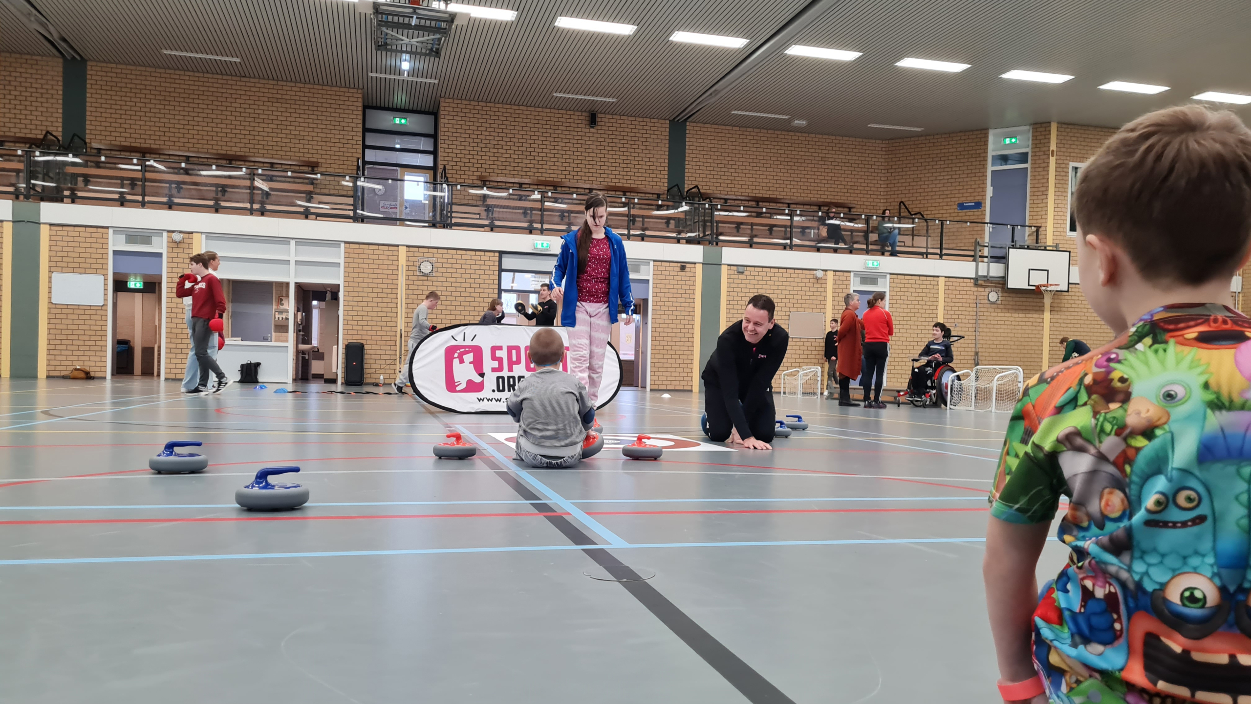 Kinderen spelen een veldcurling spel in een sporthal; enkele volwassenen zijn ook aanwezig, waaronder een volwassen begeleider die hurkt naast een kind dat op de grond zit.