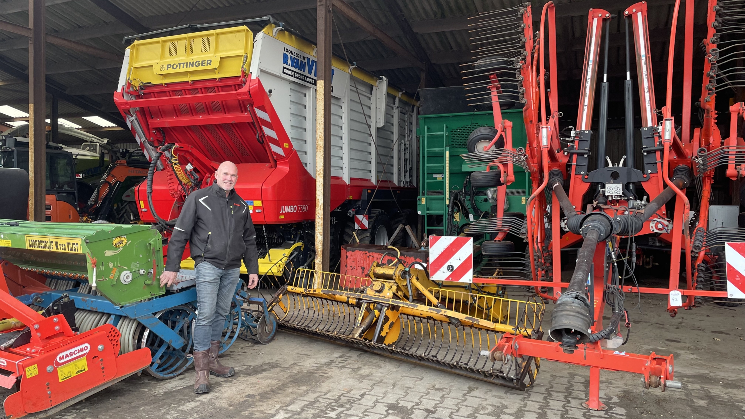 Een man staat in een garage vol landbouwmachines, waaronder een grote rode Pöttinger machine, omgeven door groene en rode landbouwapparatuur op een betonnen vloer.