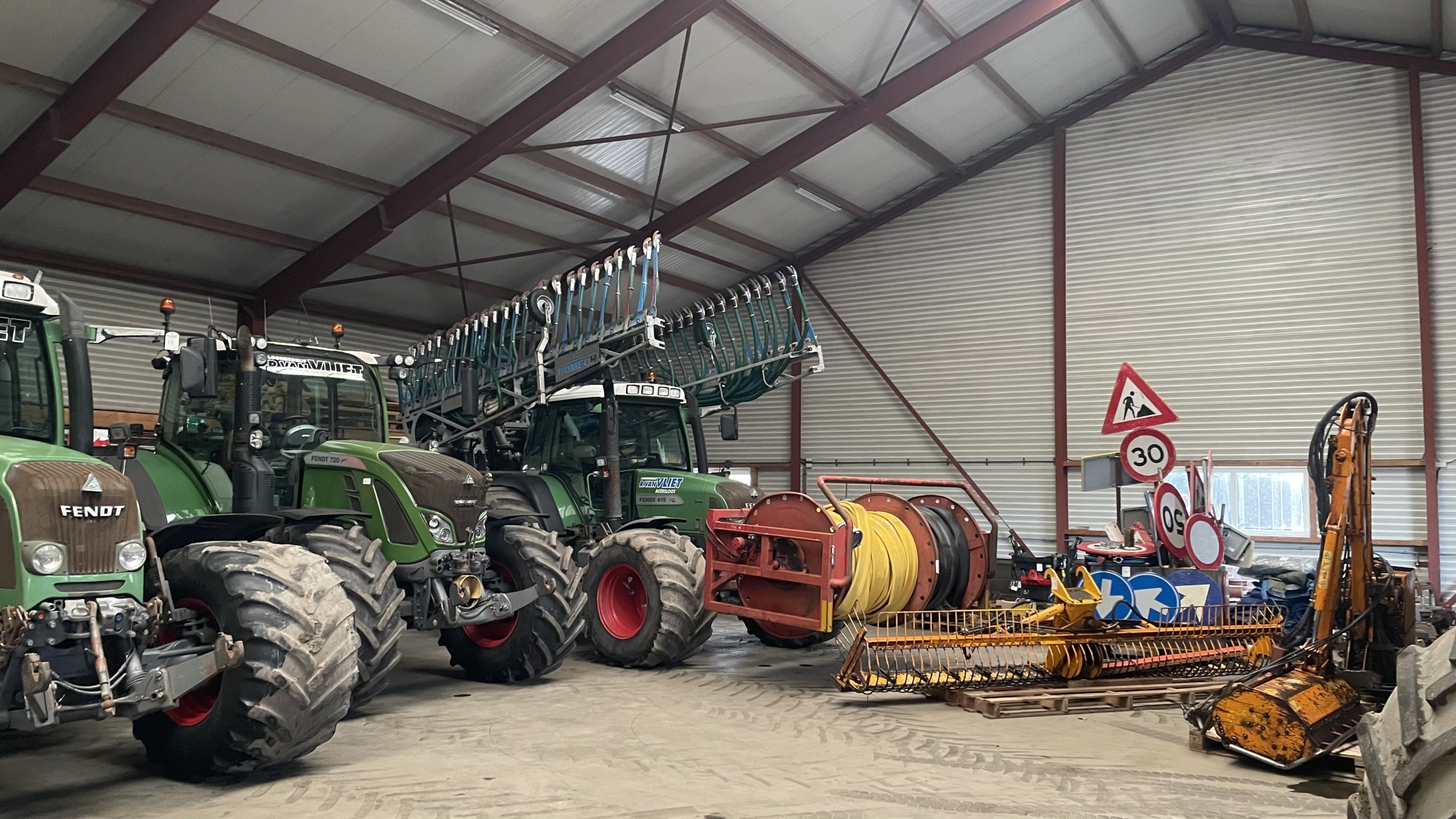 Een schuur met grote groene trekkers van het merk Fendt, een landbouwspuit, diverse verkeersborden en andere landbouwapparatuur.