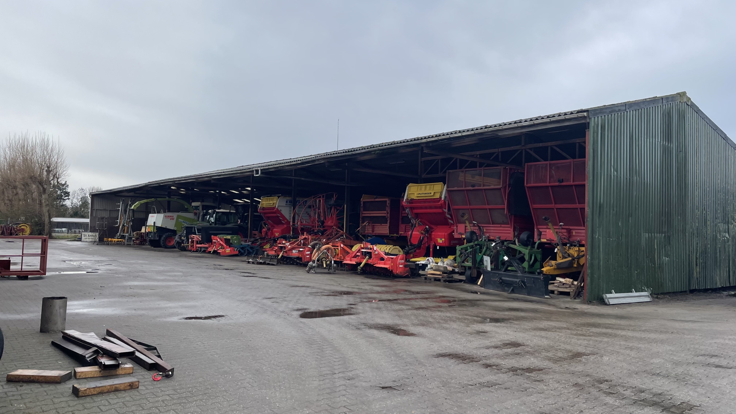 Een open loods met verschillende landbouwmachines en apparatuur, waaronder grote rode en groene voertuigen, op een bewolkte dag.