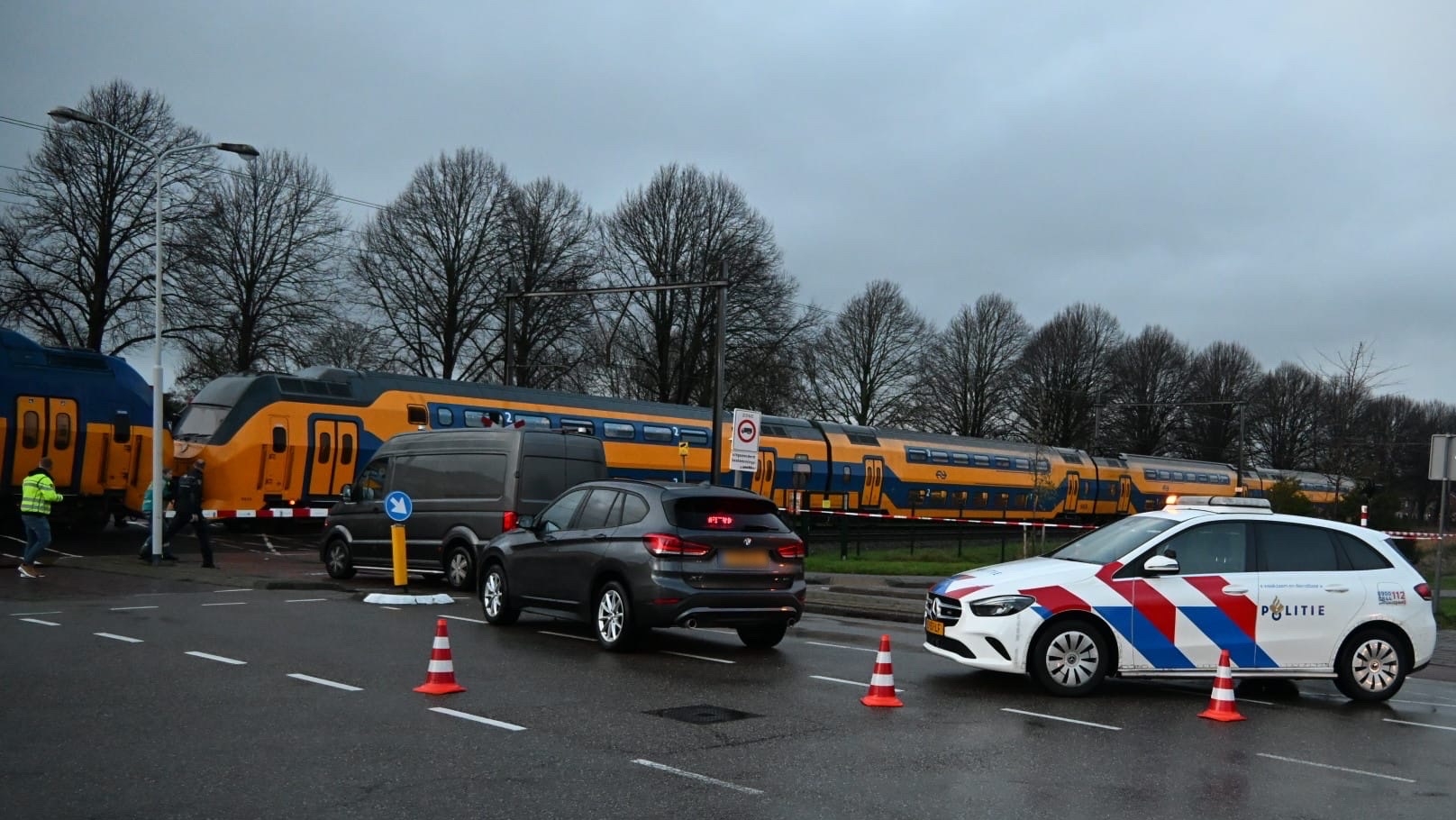 Een politieauto en andere voertuigen bij een wegafzetting voor een passerende dubbeldekstrein, met enkele mensen op de achtergrond.