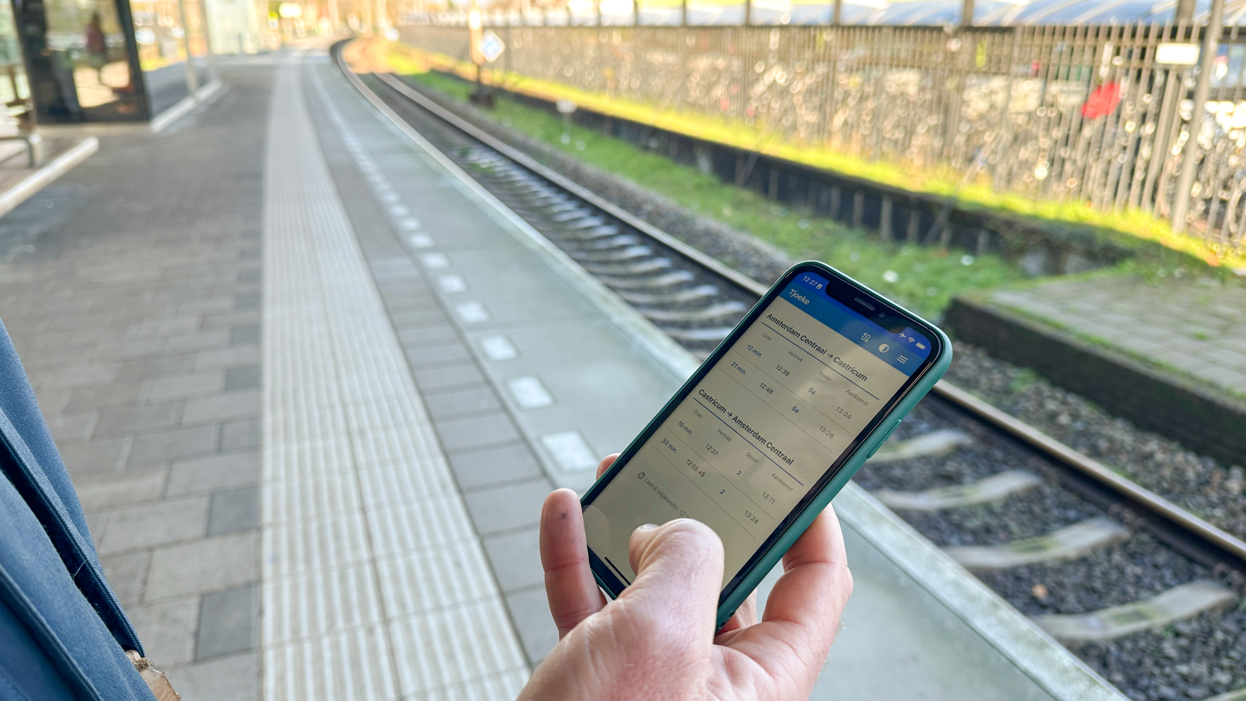 Een persoon houdt een smartphone met een reisinformatie-app vast op een leeg treinstation met spoorrails op de achtergrond.