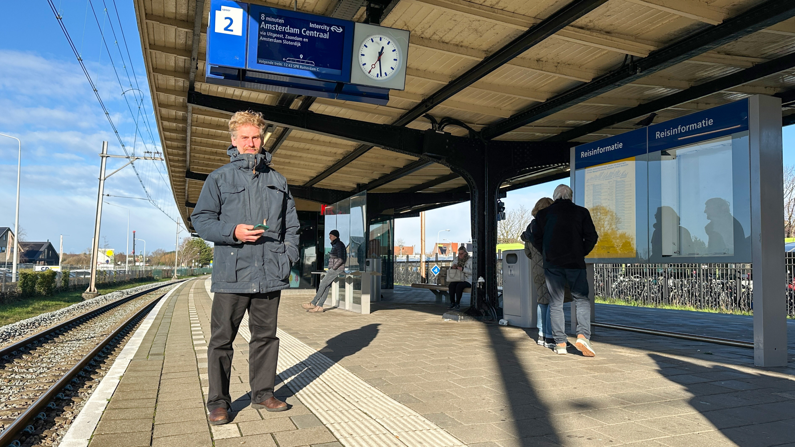Een man staat op een perron bij station Castricum, Nederland, terwijl hij een mobiel apparaat vasthoudt. Er hangen borden met reisinformatie en een klok boven het perron. Enkele andere mensen staan en zitten in de buurt van de overkapping. De lucht is helder en het is zonnig.