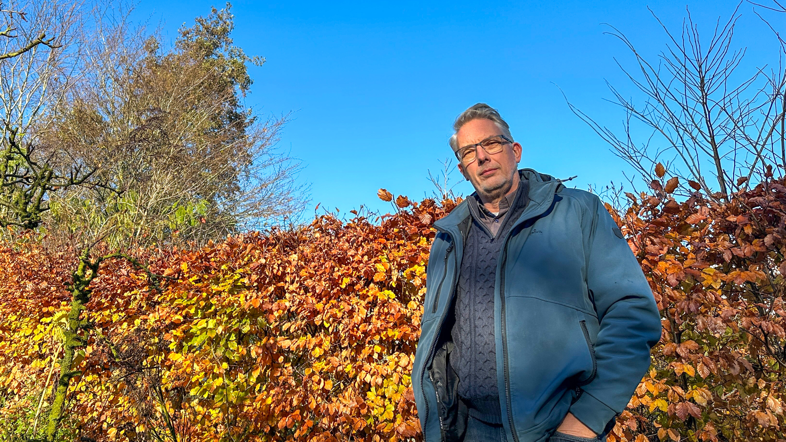 Man met bril en blauw jack staat voor een haag met herfstbladeren onder een blauwe hemel.