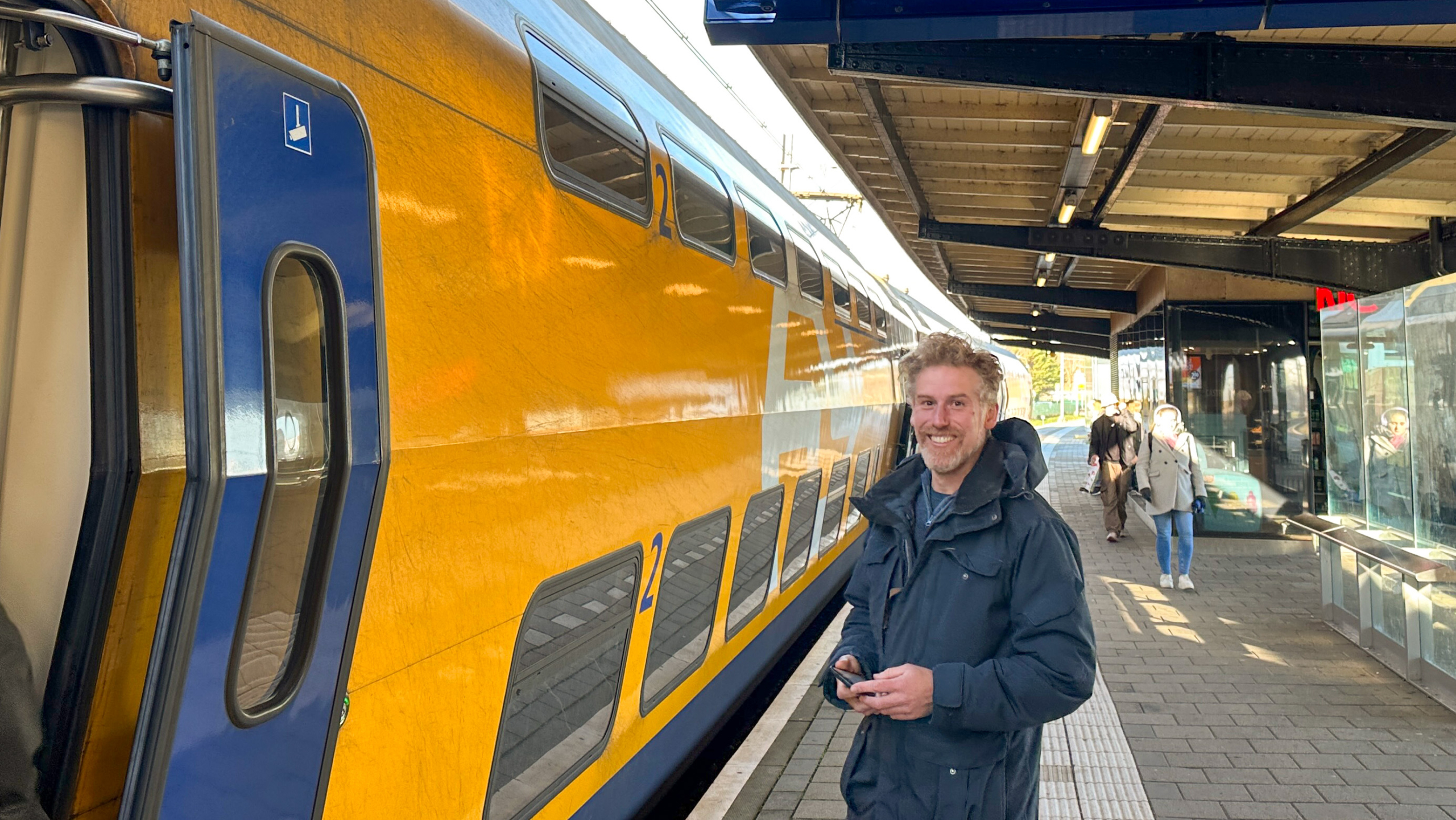 Een man glimlacht bij een gele NS dubbeldekker trein op een perron.