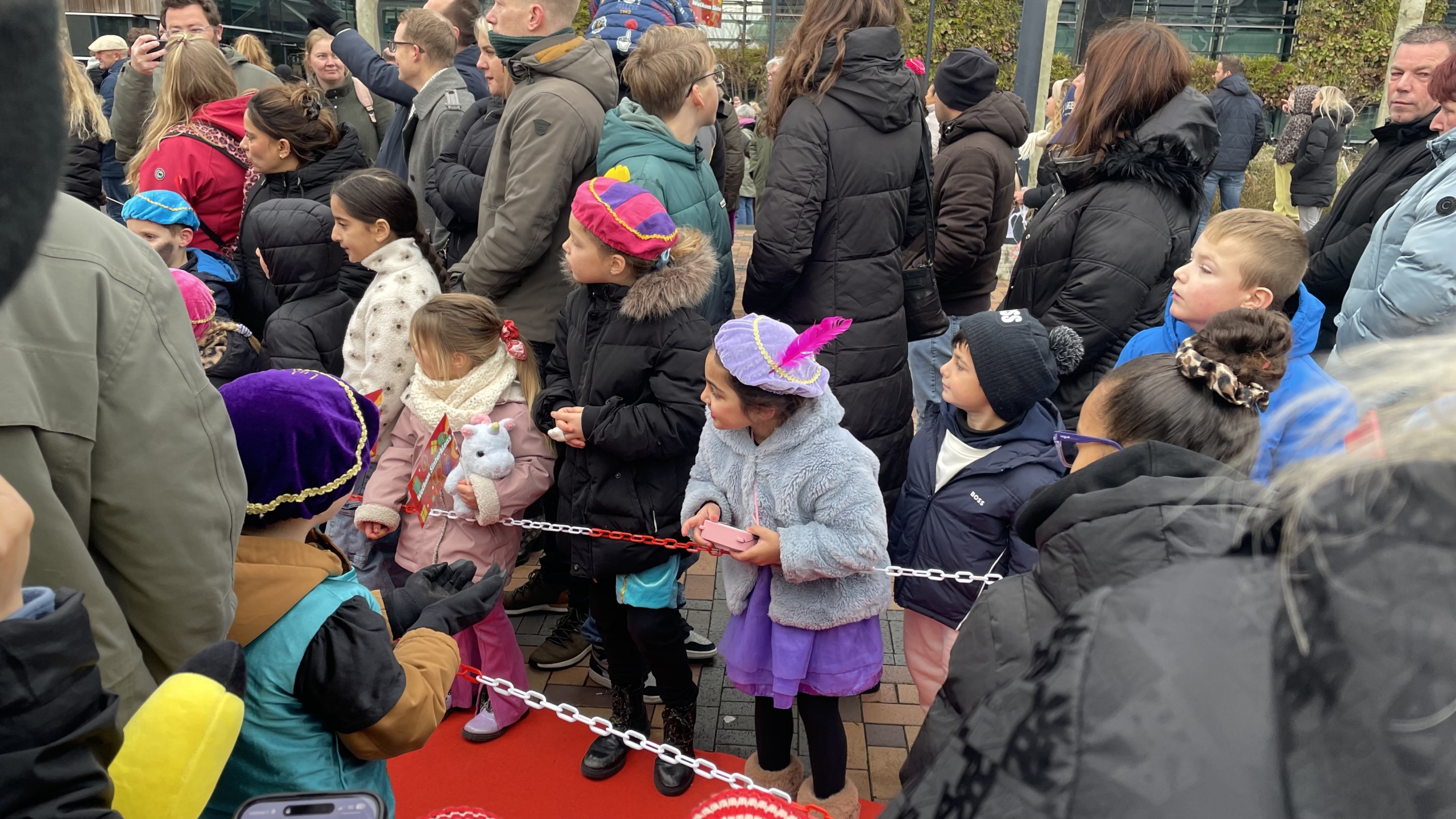 Kinderen in warme kleding en kleurrijke hoeden staan achter een afzetting tijdens een buitenbijeenkomst, omringd door een groep volwassenen.