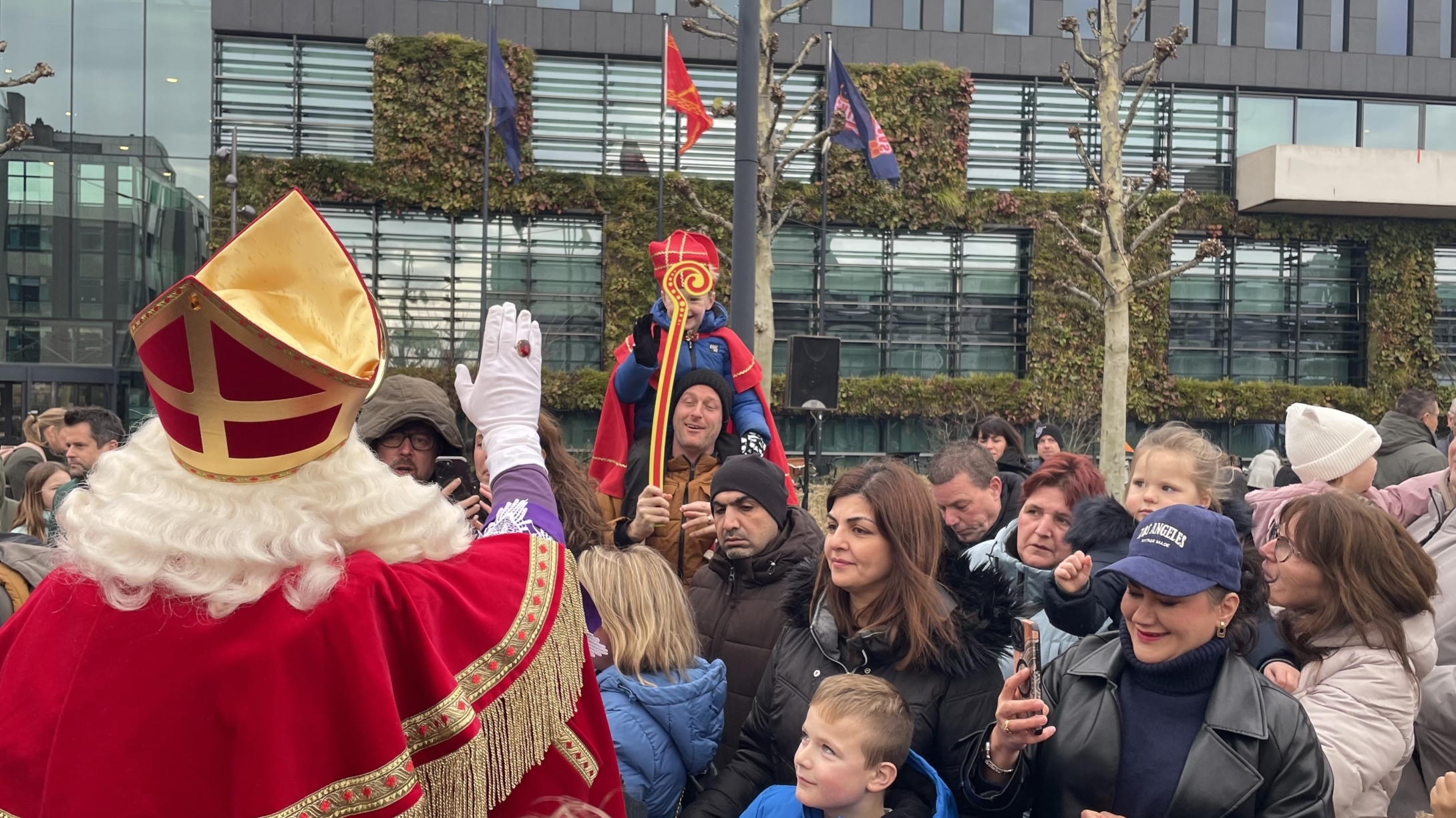 Groep mensen verzamelt zich rond een figuur gekleed als Sinterklaas, met een rode mantel en mijter, in een stadspleinomgeving. Kinderen en ouders lijken enthousiast terwijl ze naar de figuur kijken. Een mengeling van moderne gebouwen is zichtbaar op de achtergrond.
