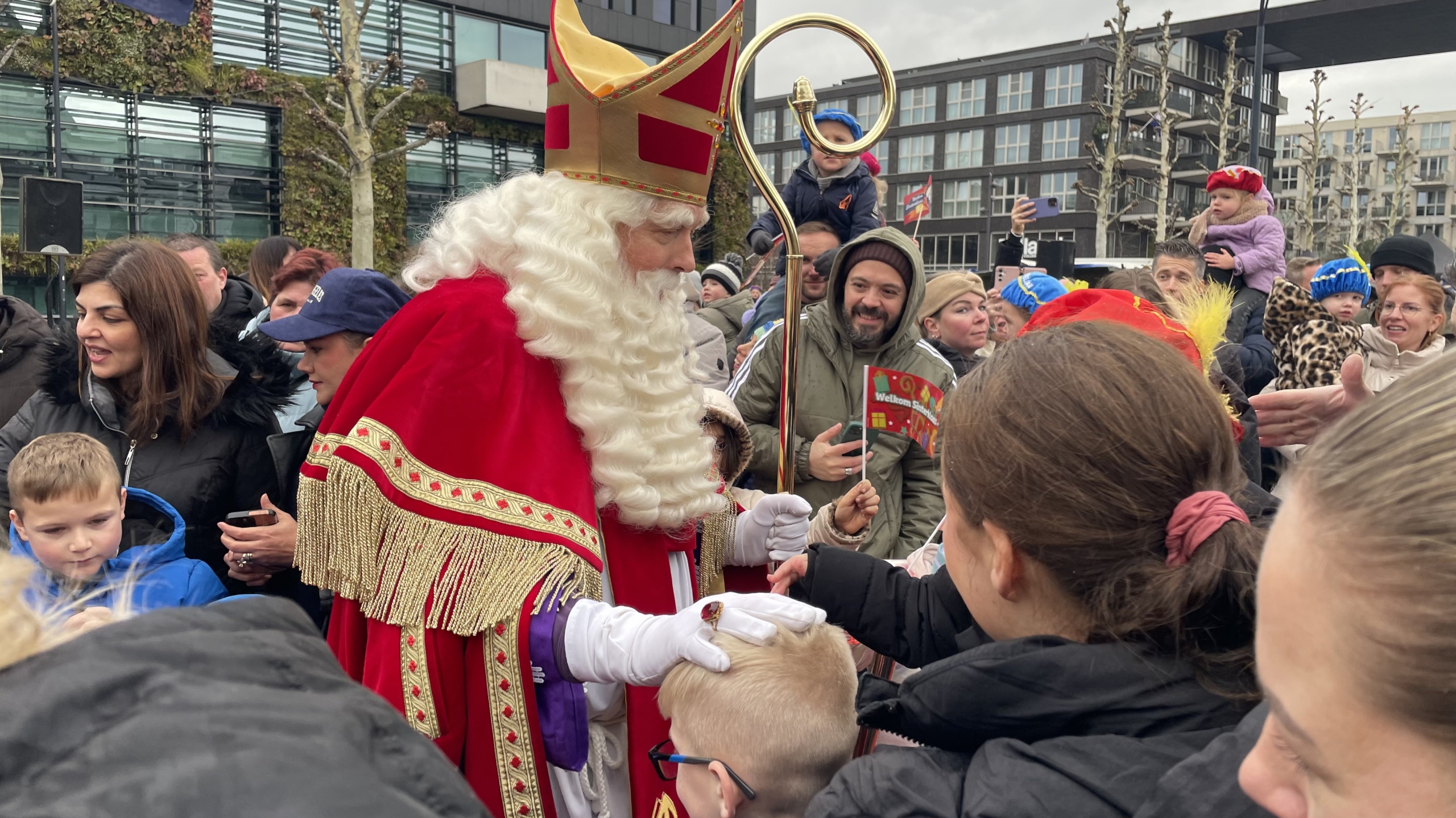Sinterklaas omringd door een menigte, geeft een kind een hand in een feestelijke buitenomgeving met mensen die hem fotograferen.