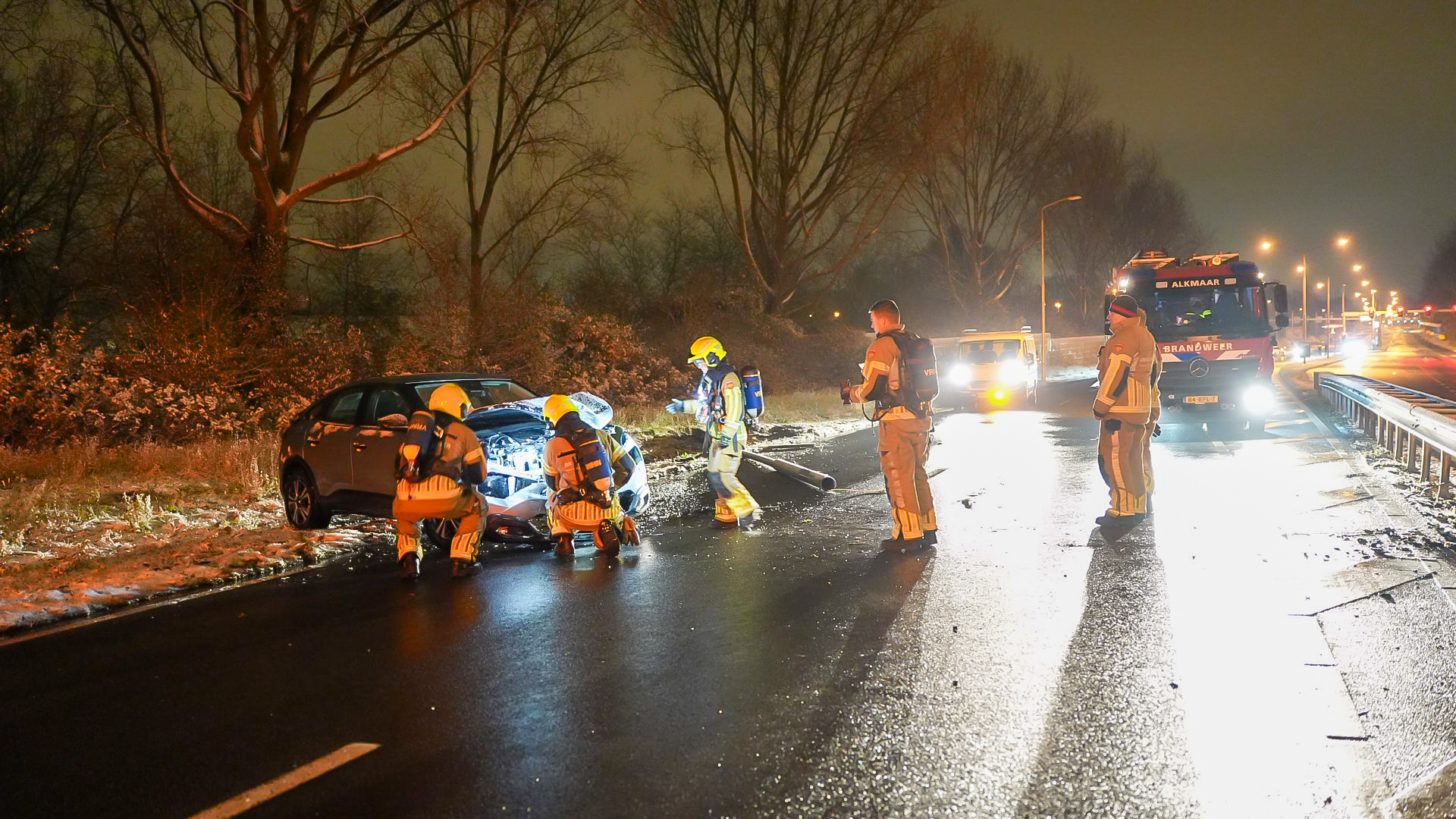Brandweerlieden onderzoeken een auto na een ongeval op een natte weg bij nacht, met een brandweerwagen op de achtergrond.