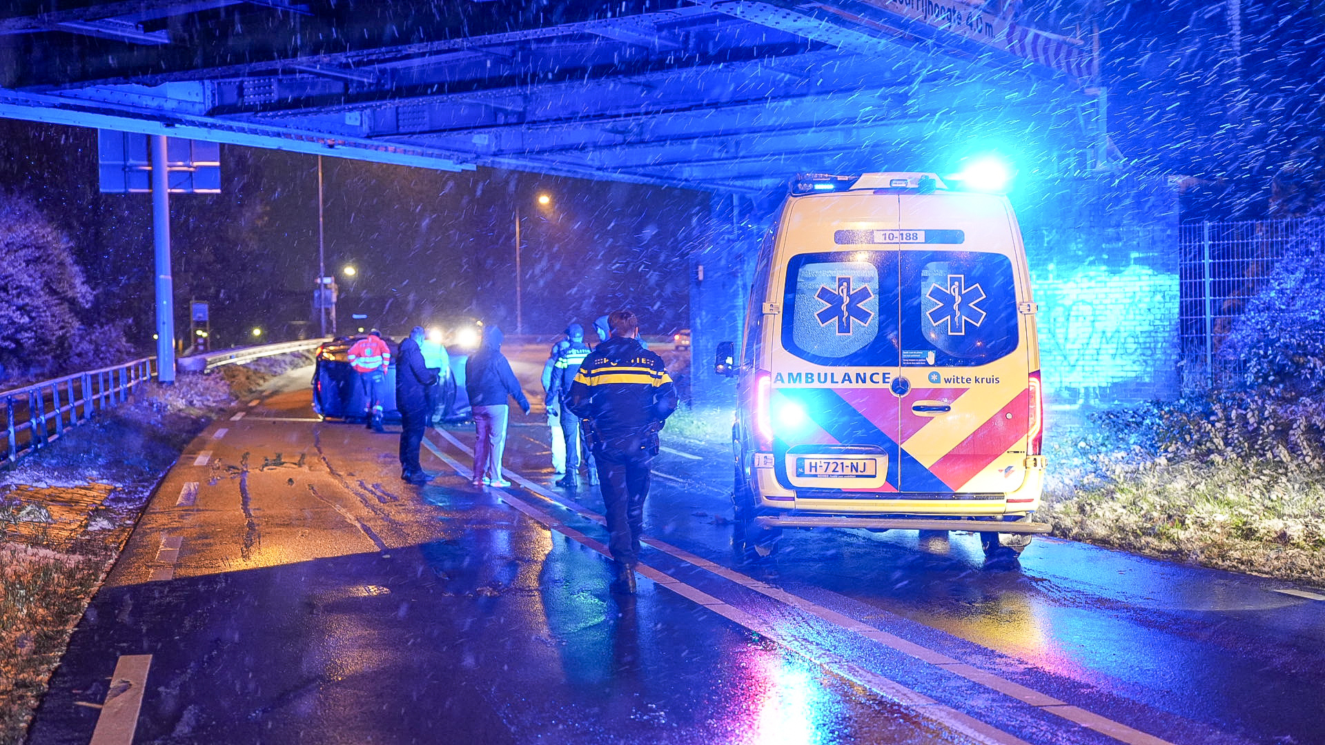 Ambulance en politie arriveren bij een ongeval op een besneeuwde weg onder een viaduct, 's nachts met blauwe zwaailichten.