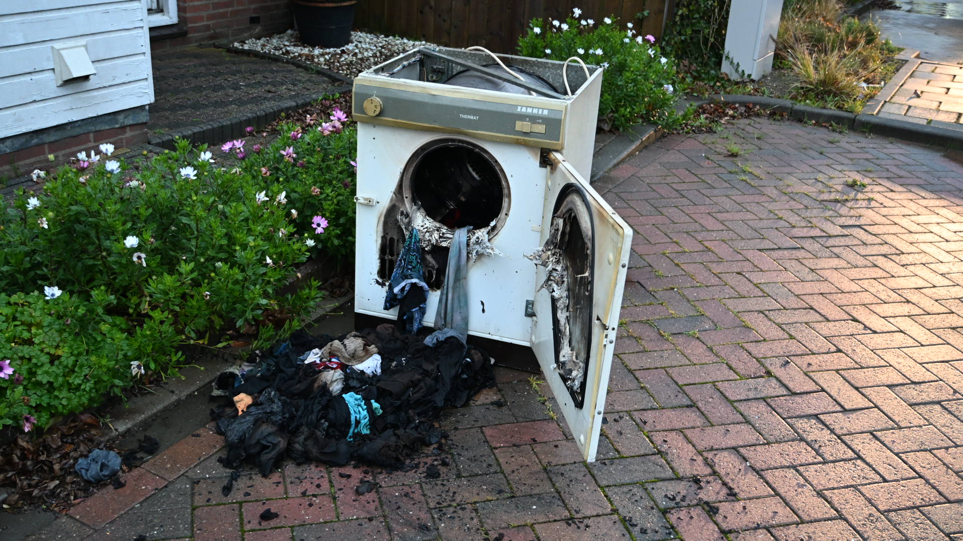 Een kapotte wasmachine met beschadigde kleding ervoor, geplaatst op een betegeld pad naast een bloeiende tuin, onder een avondzonlicht.