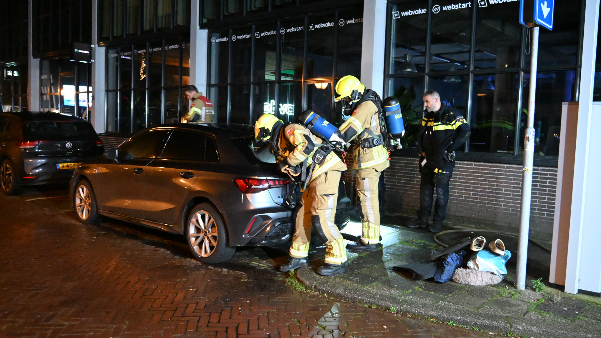 Brandweerlieden onderzoeken een geparkeerde auto 's nachts in een stedelijke omgeving, omstanders en politieman in de buurt.