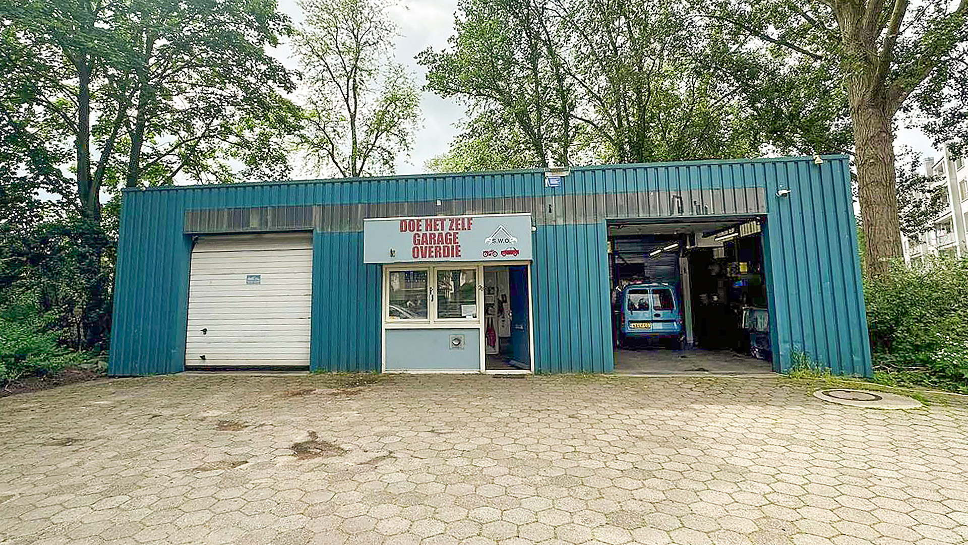 Blauwe garage met opschrift 'DOE HET ZELF GARAGE OVERDIE', gelegen in een groen, bosrijk gebied met een bestelwagen geparkeerd in de geopende garage.