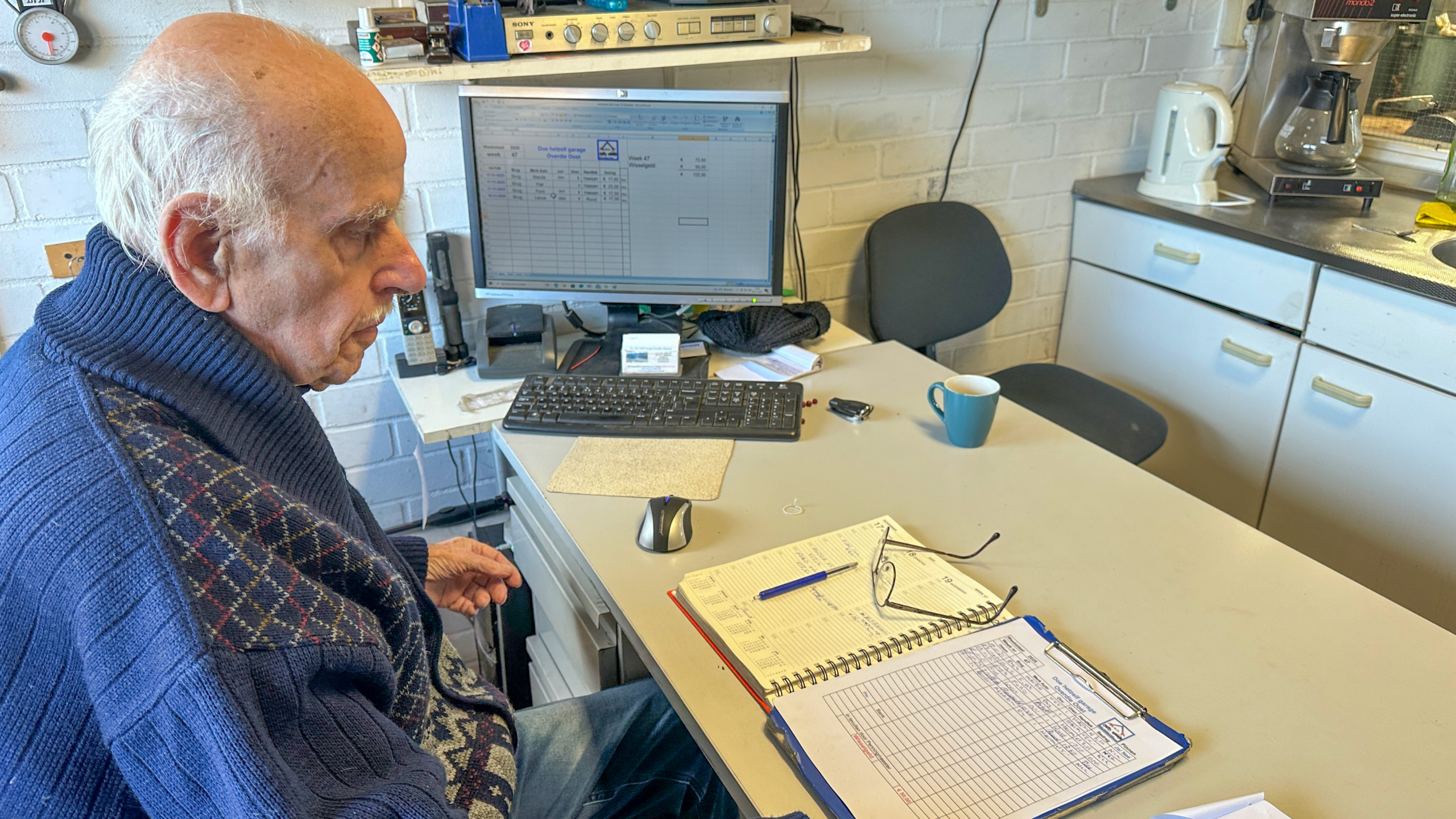 Oude man zittend aan een bureau met een computer, schrijfmateriaal en een mok op tafel, in een kantooromgeving met witte bakstenen muren en een koffiezetapparaat op de achtergrond.