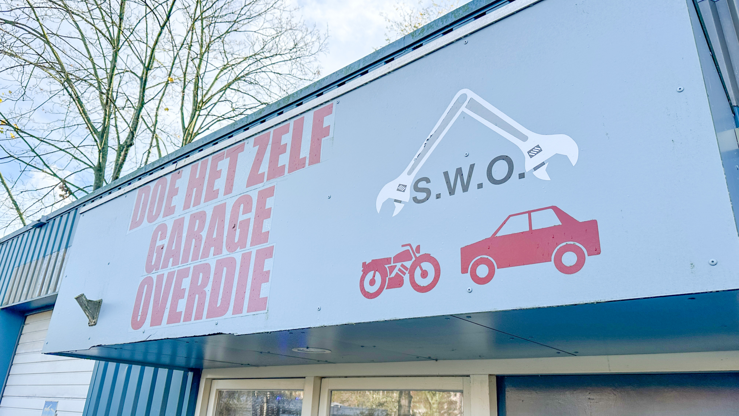 Uithangbord van "Doe het zelf Garage Overdie" met afbeeldingen van een moersleutel, een motorfiets en een auto op een wolkenloze dag.