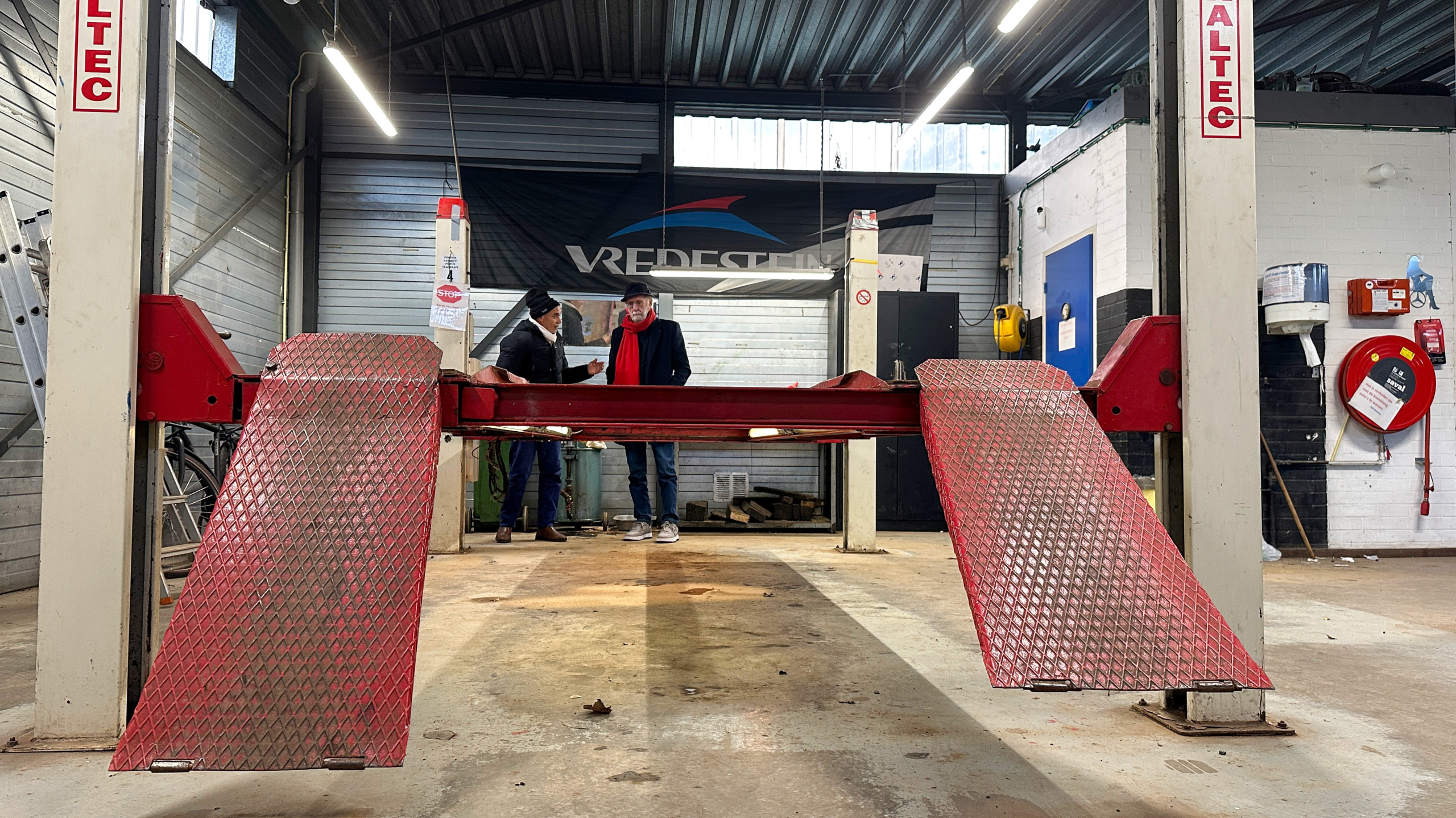 Twee mensen staan in een autowerkplaats naast een rode autolift van het merk Altec, met een Vredestein bord op de achtergrond.