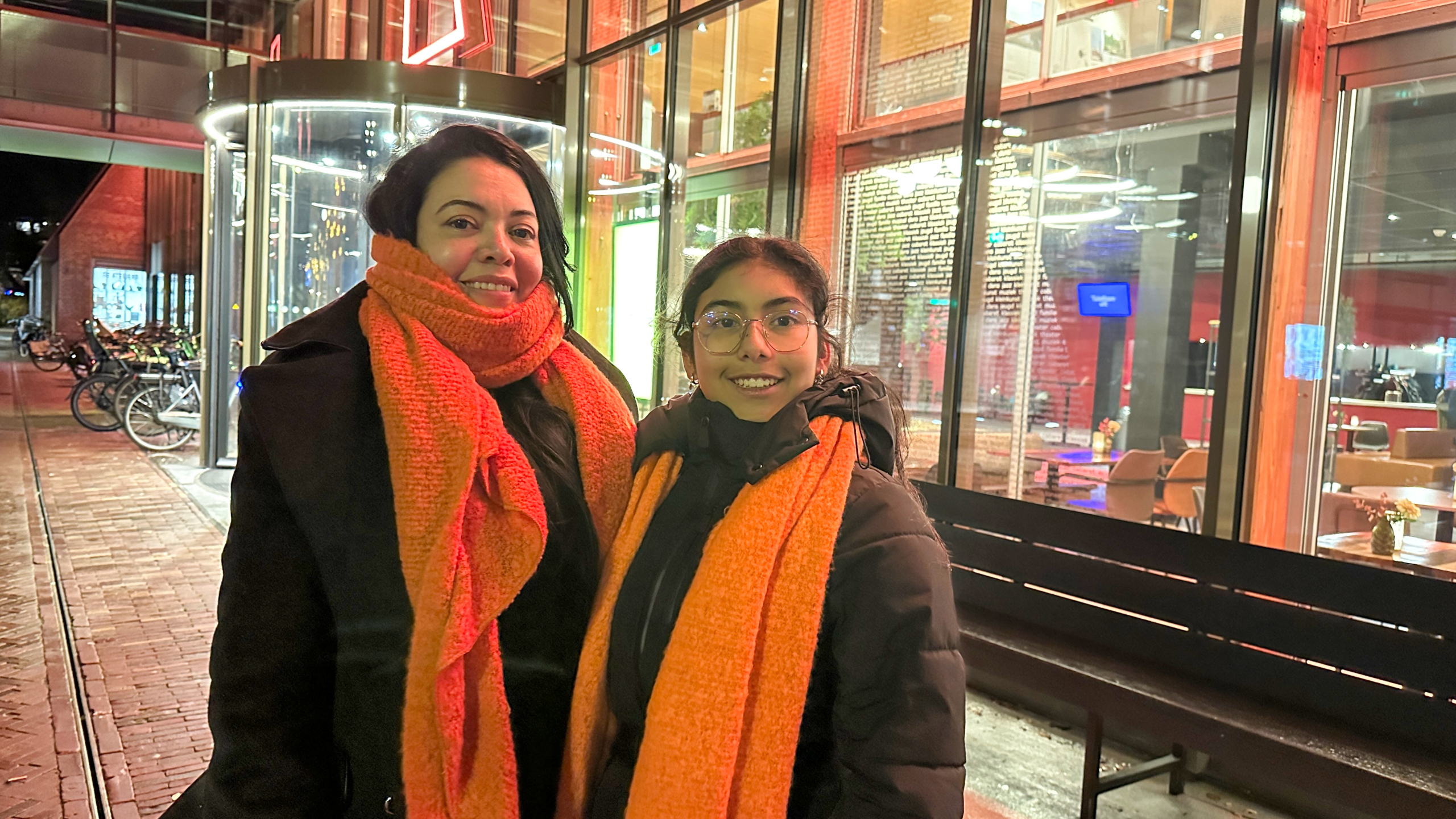 Twee mensen poseren buiten voor een glazen gebouw verlicht met rood en groen, beiden dragen zwarte jassen en oranje sjaals.