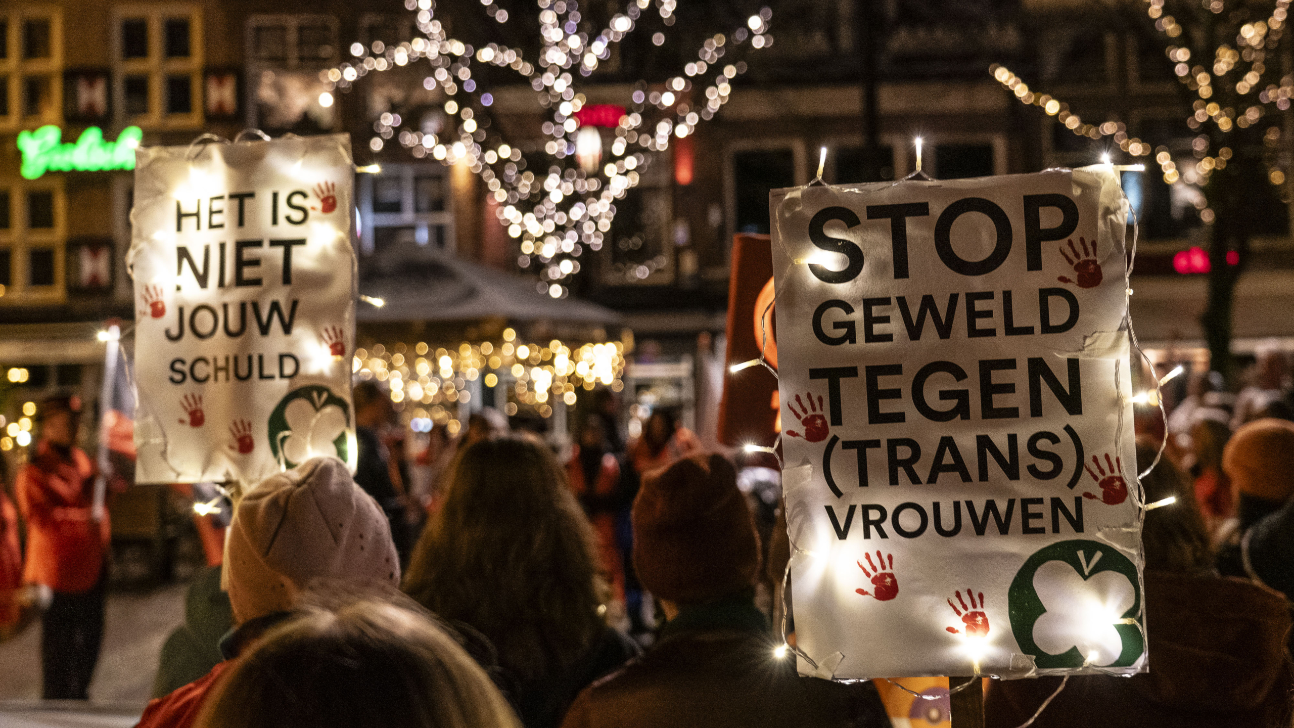 Mensen houden verlichte borden vast met de teksten 'HET IS NIET JOUW SCHULD' en 'STOP GEWELD TEGEN (TRANS) VROUWEN' tijdens een nachtelijke demonstratie met fonkelende lichtjes in de bomen op de achtergrond.