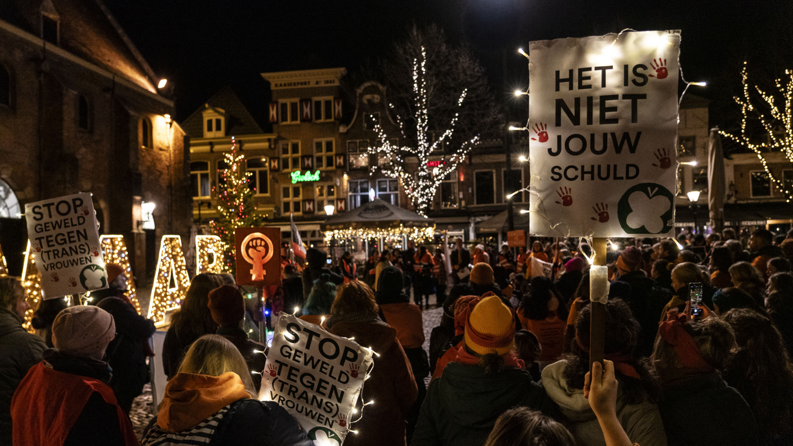 Groep mensen verzameld voor een nachtelijke demonstratie met borden tegen geweld, versierde kerstboom en feestverlichting op de achtergrond. 