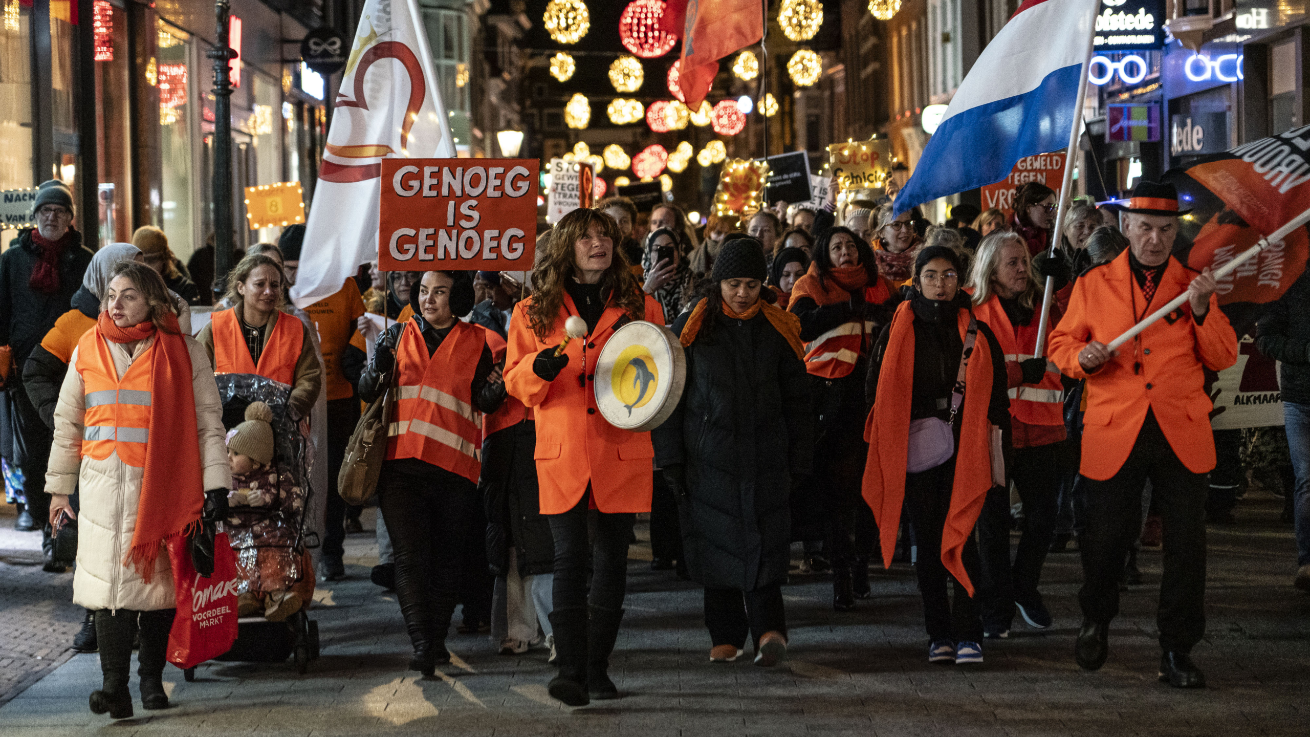 Mensen in oranje veiligheidsvesten marcheren met protestborden en vlaggen tijdens een nachtdemonstratie met feestverlichting op de achtergrond.