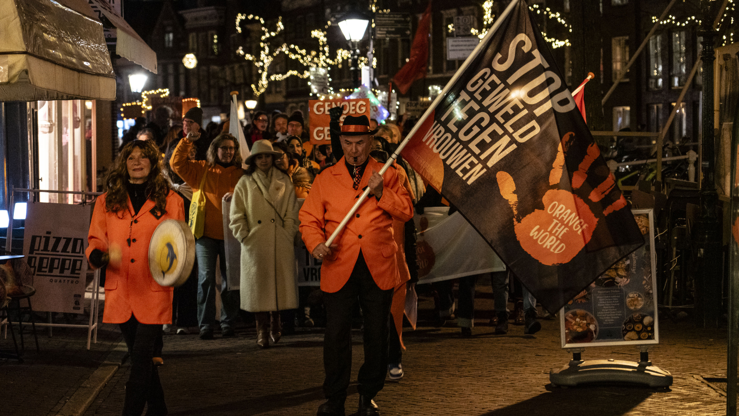 Een groep mensen loopt 's avonds in een mars met een vlag waarop staat: "Stop geweld tegen vrouwen." Zij dragen oranje kleding als onderdeel van de "Orange the World" campagne. Op de achtergrond zijn verlichte straten te zien.