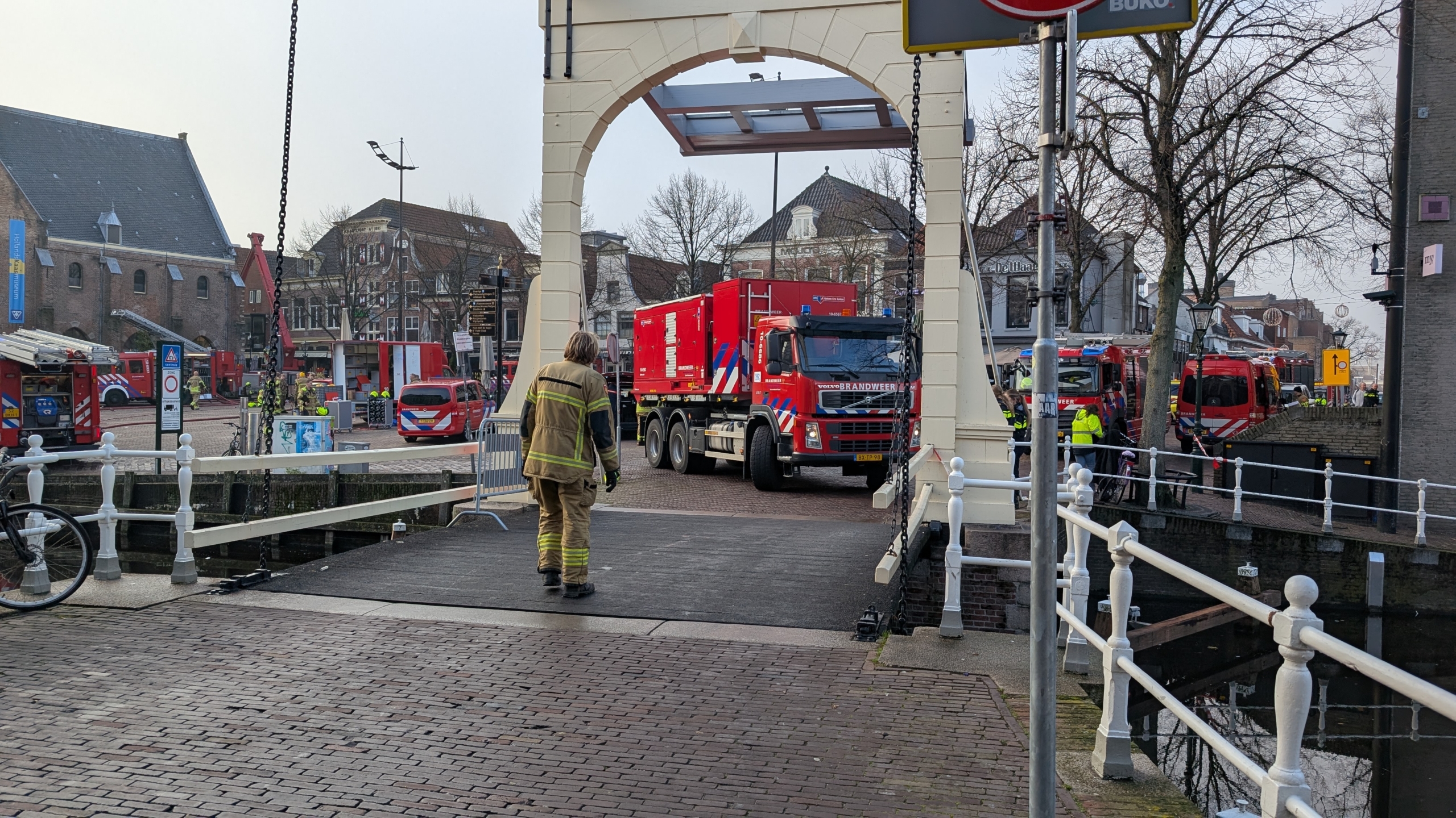 Een brandweerman loopt naar een rode brandweertruck die door een brug rijdt, omringd door meerdere brandweer- en hulpdienstvoertuigen in een stedelijke omgeving.