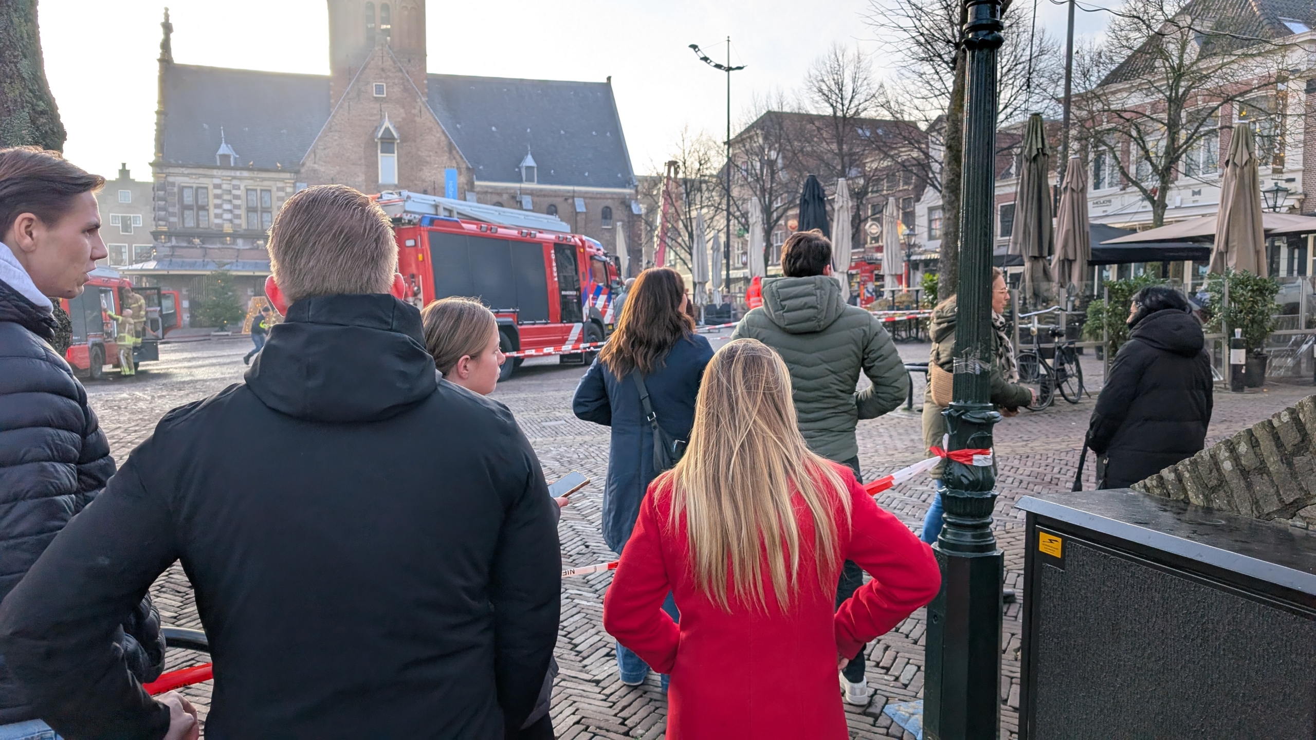 Een groep mensen staat te kijken naar een brandweerwagen op een plein, met op de achtergrond een kerk en omringende gebouwen. Het gebied is afgezet met rood-wit lint.