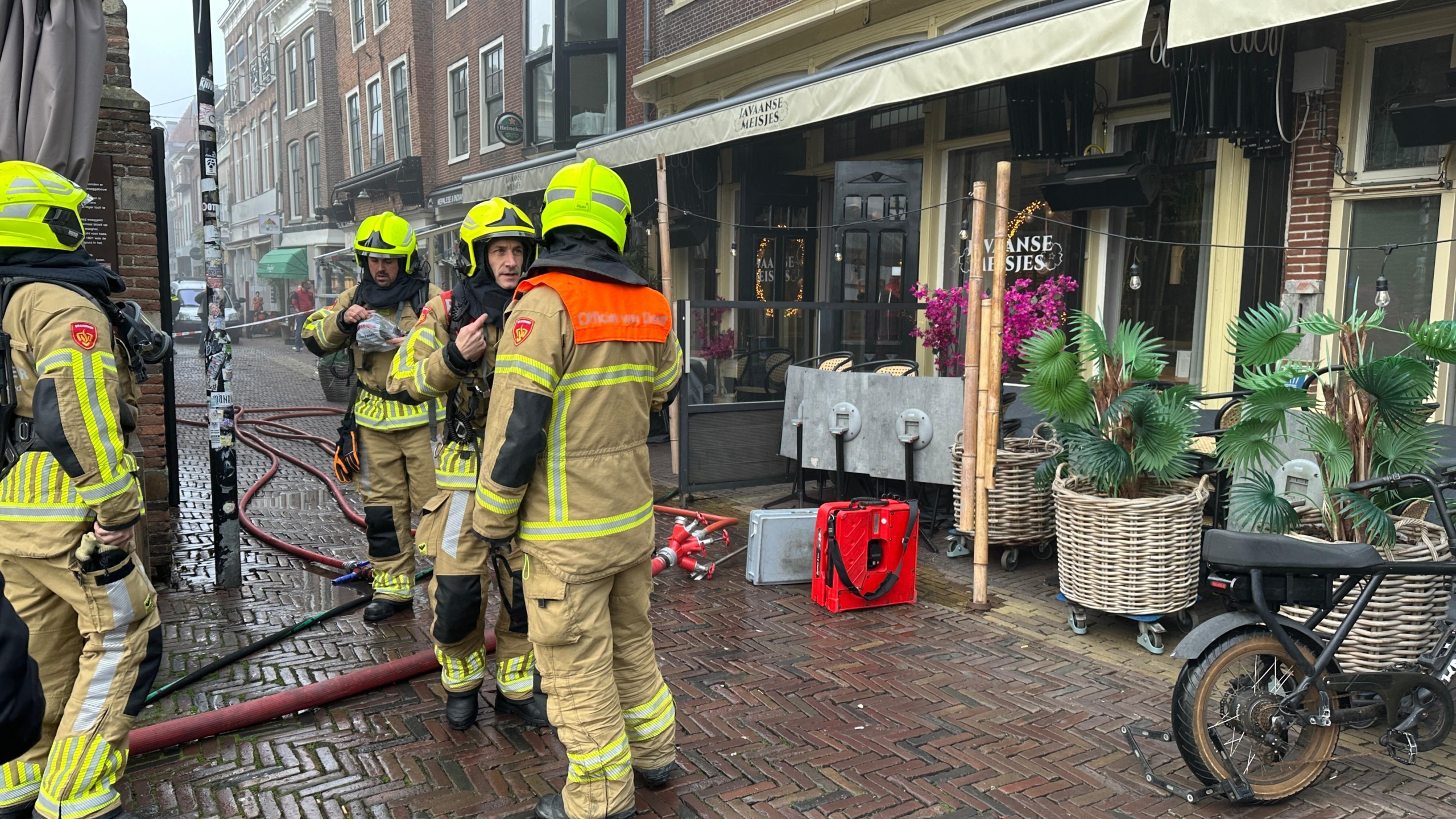 Brandweermannen in uniformen staan op een straat met natte klinkers voor een café genaamd 'Javaanse Meisjes', omgeven door slangen en reddingsmateriaal.