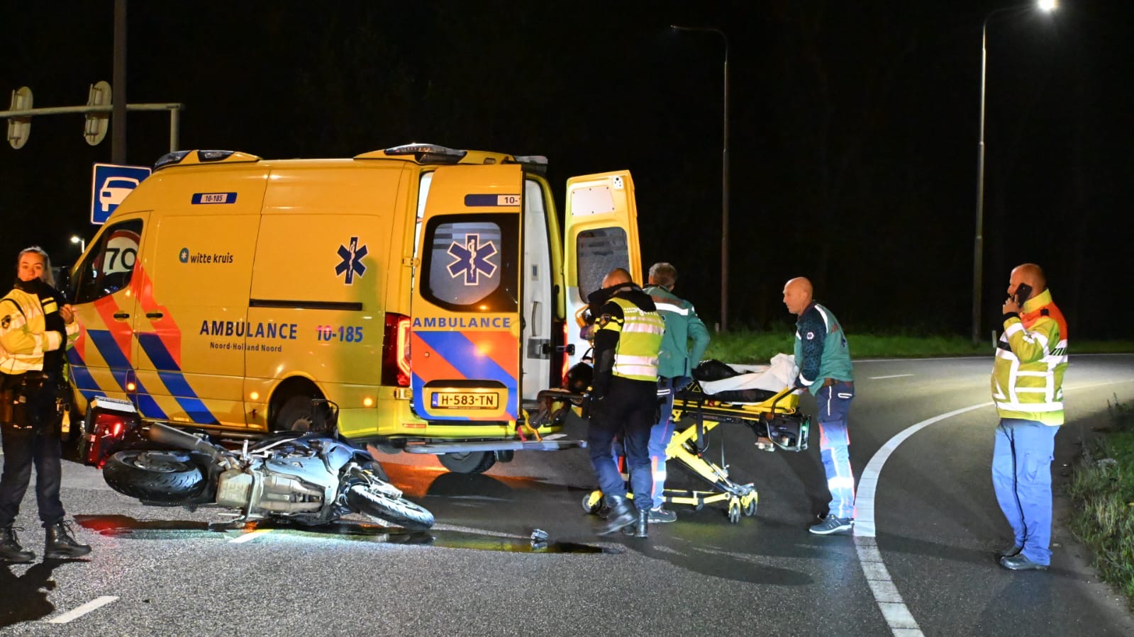 Een incident op een donkere weg met een ambulance, politieagenten, en medisch personeel die een persoon op een brancard behandelen naast een omgevallen motorfiets.