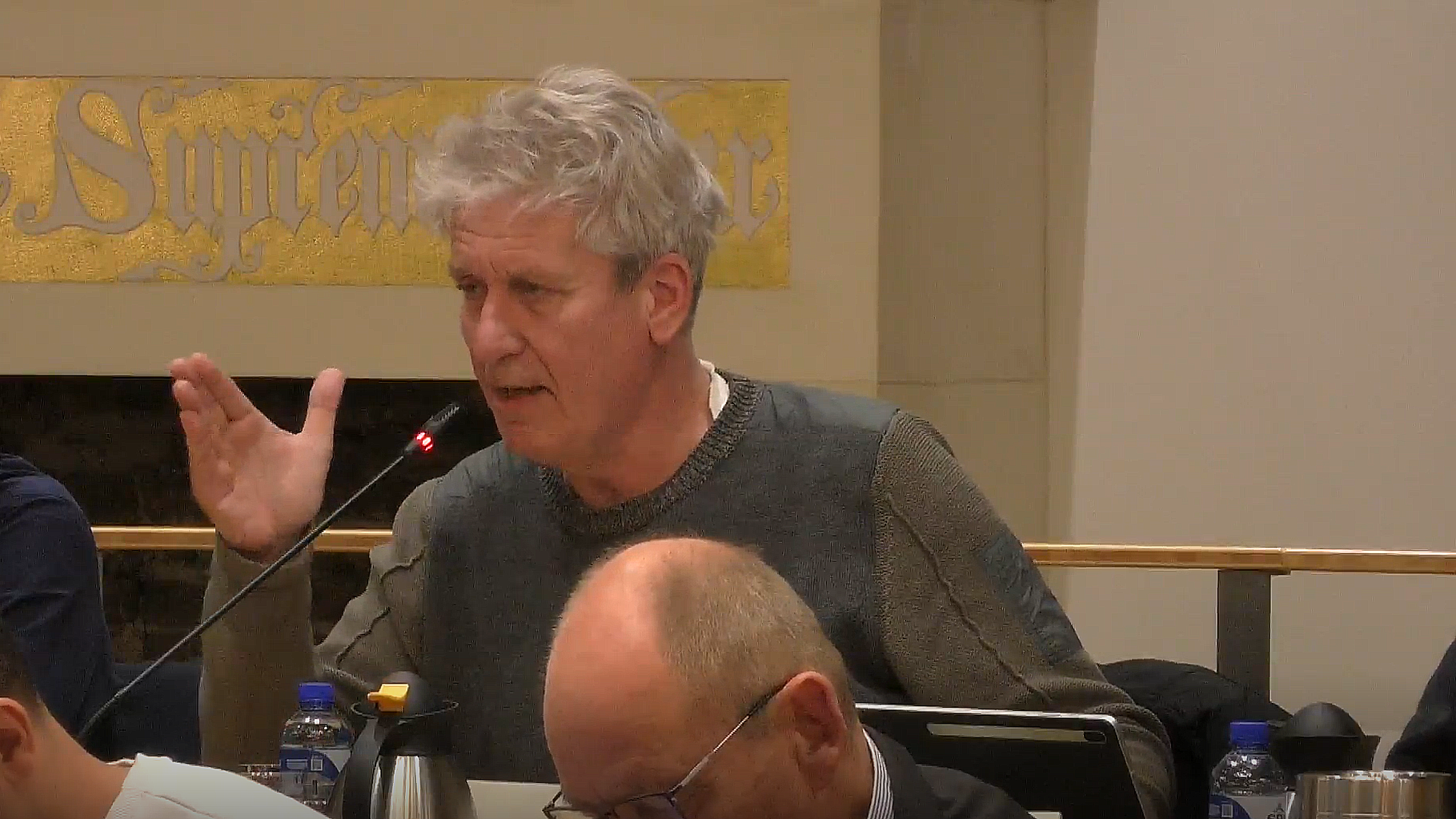 Een man met grijzend haar spreekt in een microfoon tijdens een vergadering, met een gebaar van zijn hand, terwijl een andere man, kalend, naar beneden kijkt op de voorgrond.