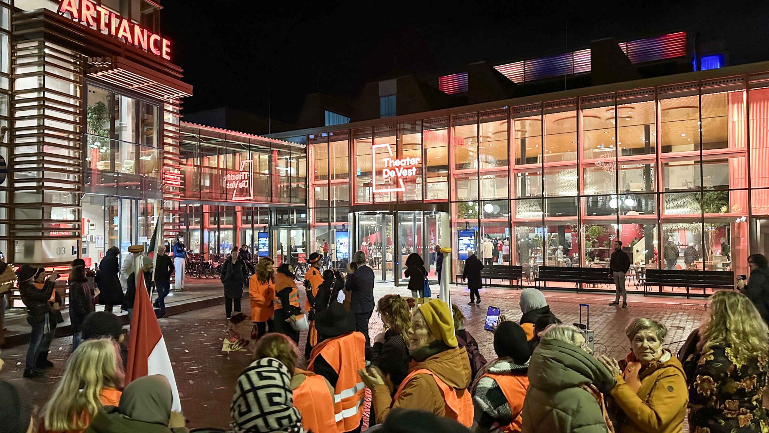 Groep mensen in oranje veiligheidshesjes voor Theater De Vest in de avond, met helder verlichte gevel en glazen ramen waarbinnen mensen zichtbaar zijn.