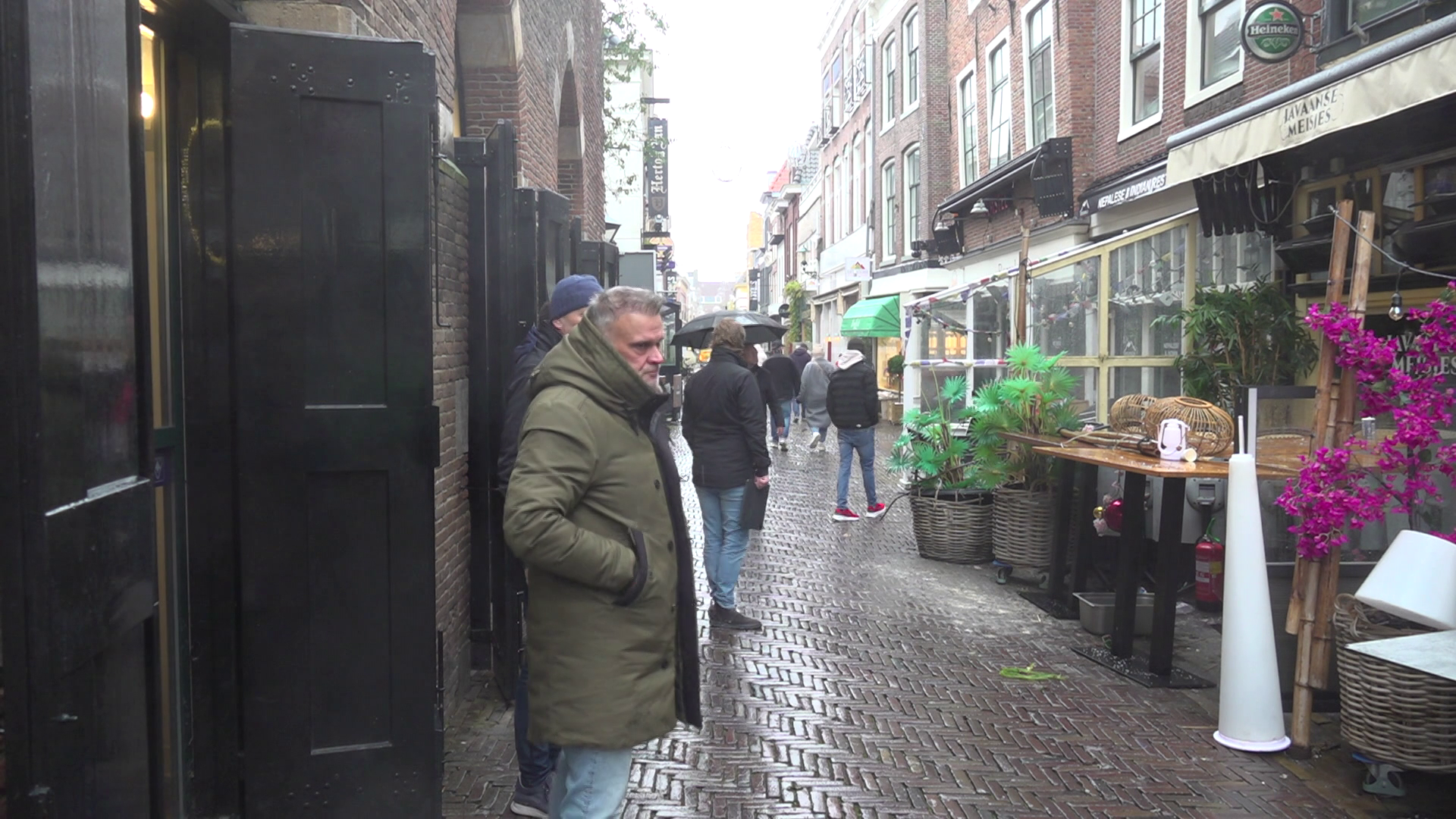 Een man in een groene jas staat op een natte, smalle straat met kasseien, omringd door mensen met paraplu's. Aan de rechterkant zijn er bloempotten en een café met borden en decoraties.
