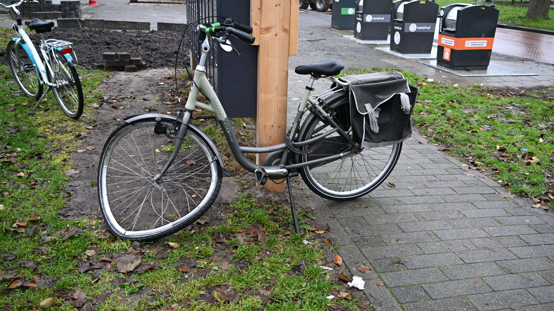 Grijze fiets met dubbele fietstas, geparkeerd tegen een houten paal op een trottoir naast een grasveldje.
