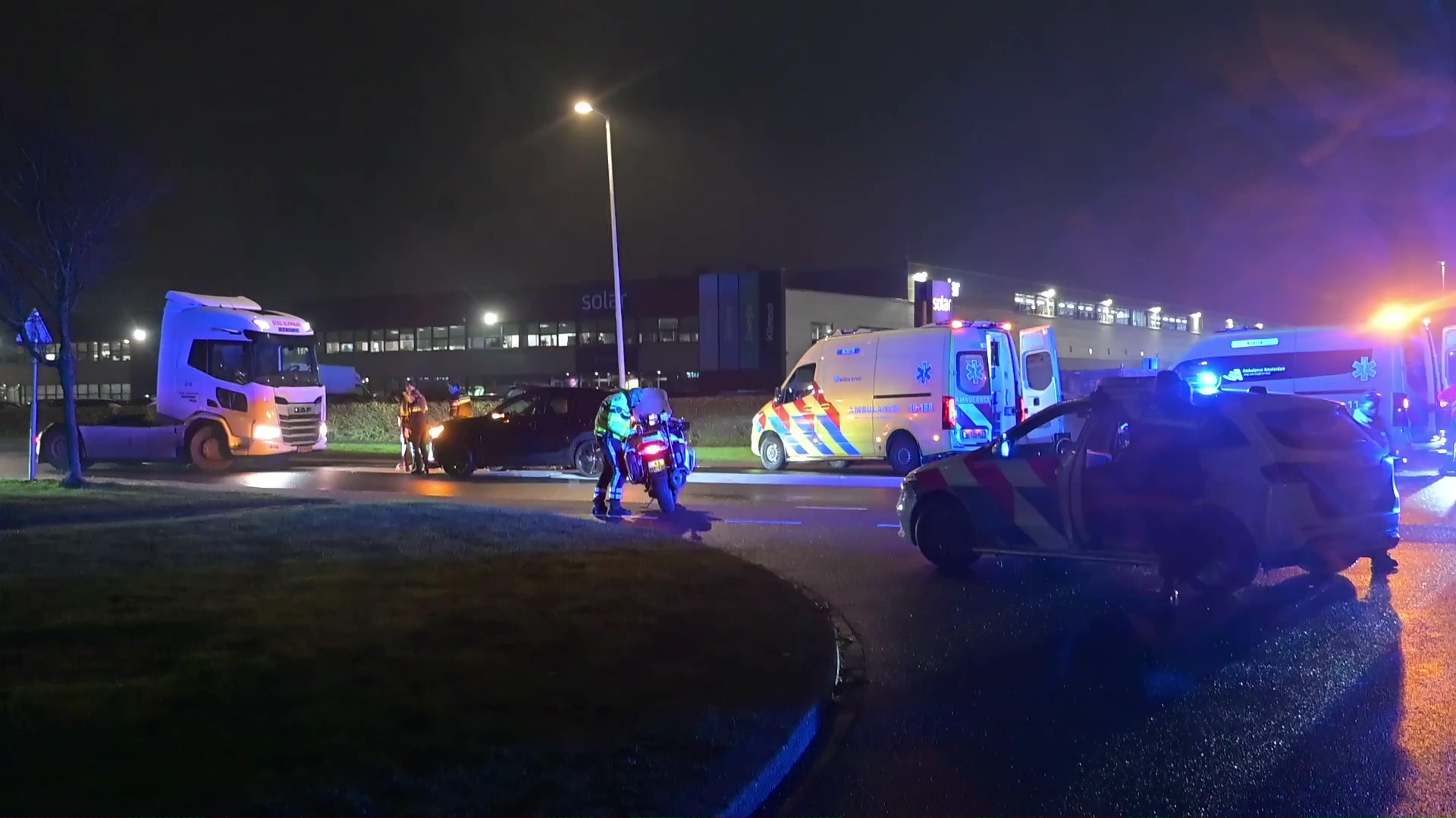 Nachtelijke scène met politie, een ambulance en een vrachtwagen op een weg, omgeven door blauwe en oranje noodverlichting.