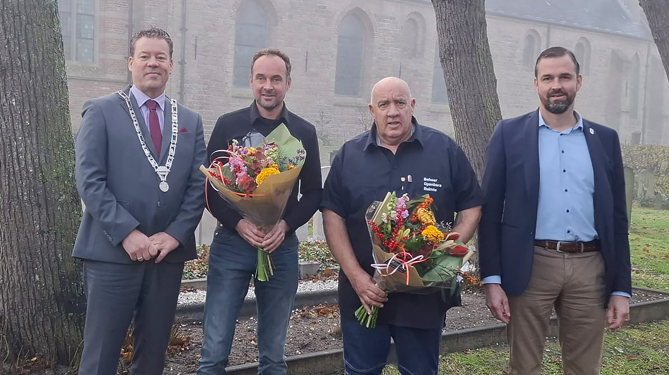 Vier mannen poseren buiten voor een kerkgebouw, twee van hen houden bloemenboeketten vast, omringd door grafstenen op de achtergrond.