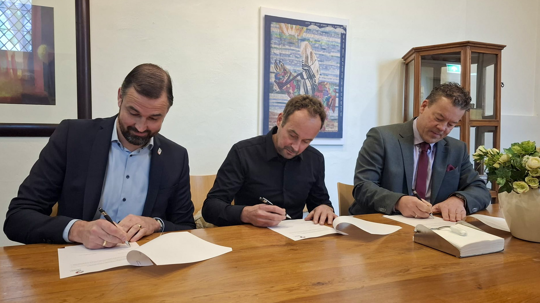 Drie mannen in zakelijke kleding ondertekenen documenten aan een houten tafel in een kantoorruimte met kunst aan de muur en bloemen op tafel.