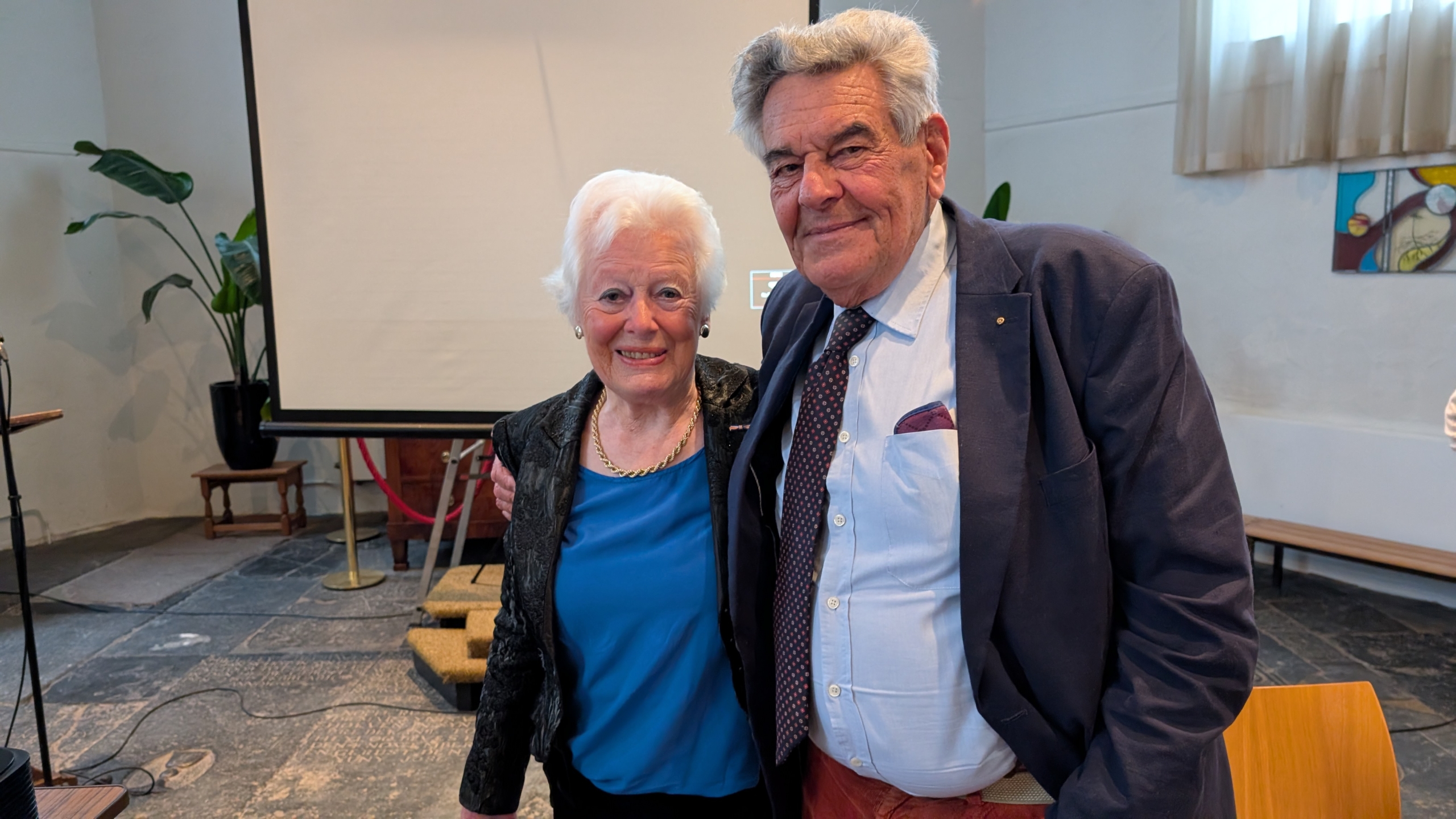 Een oudere man en vrouw glimlachen samen naar de camera in een zaal met een projectiescherm en planten op de achtergrond.