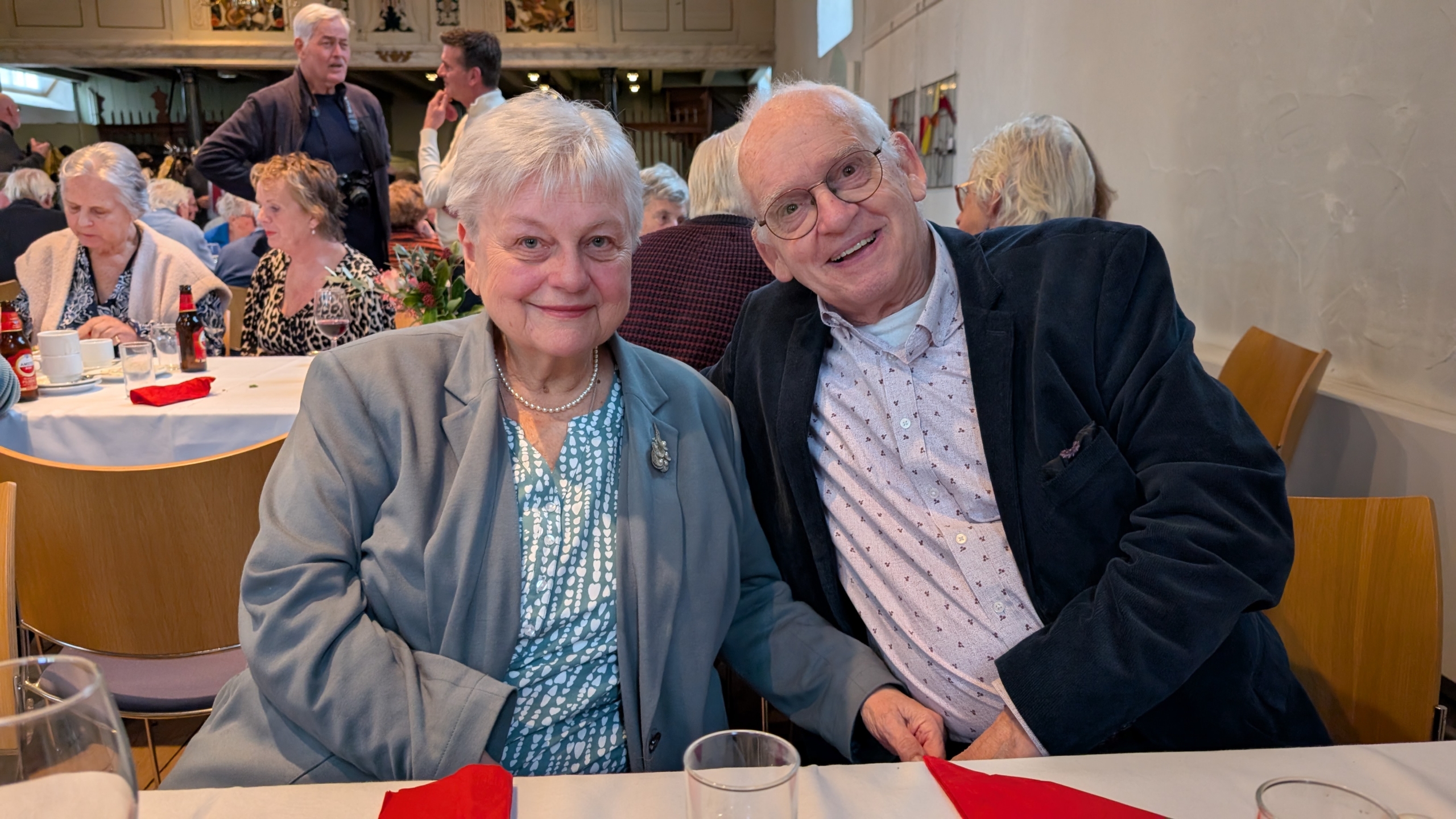 Een oudere man en vrouw zitten glimlachend aan een tafel tijdens een bijeenkomst, omringd door andere etende en pratende gasten in een zaal.