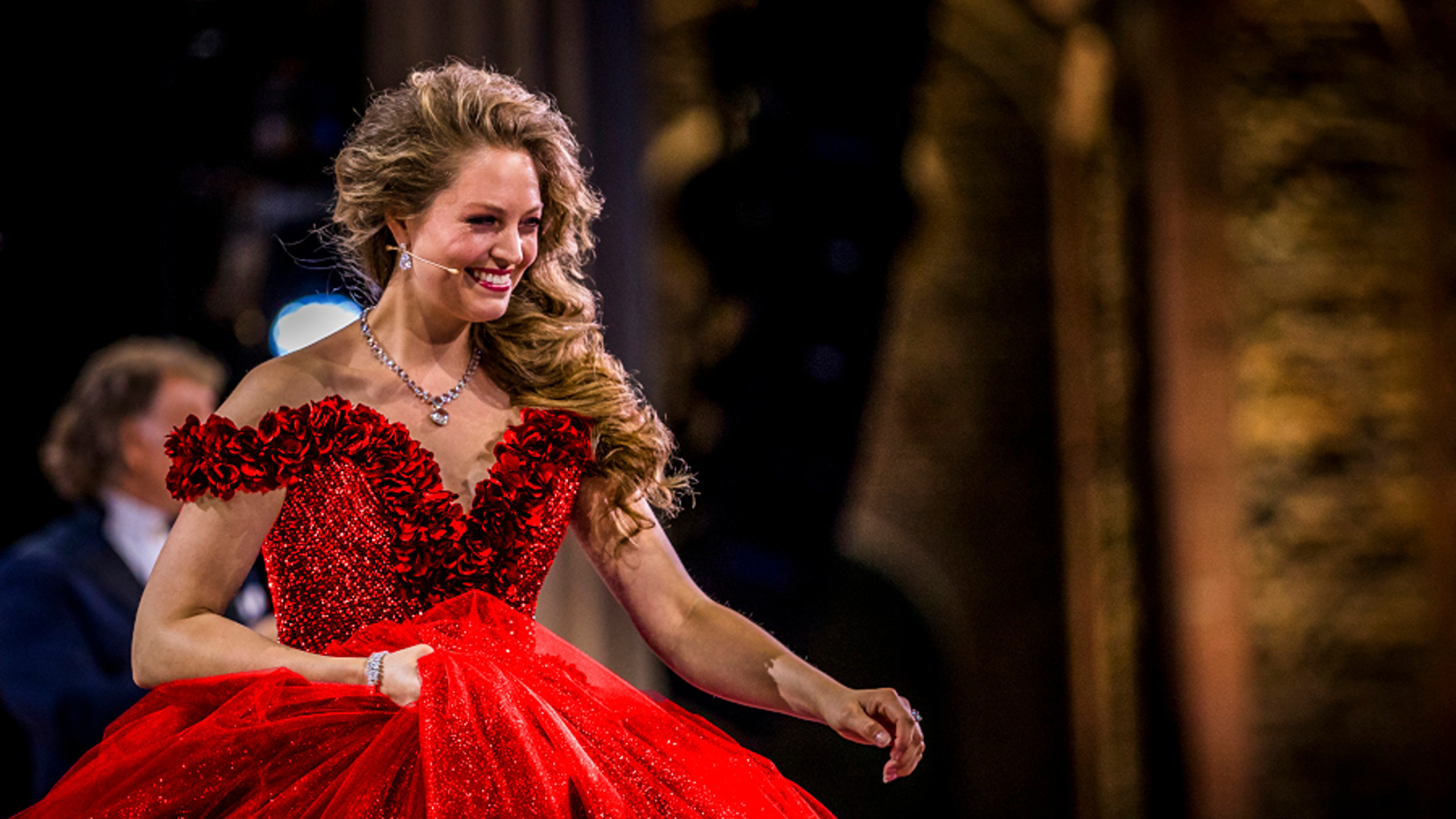 Een vrouw in een opvallende rode avondjurk met bloemenaccenten glimlacht en loopt over het podium tijdens een evenement.
