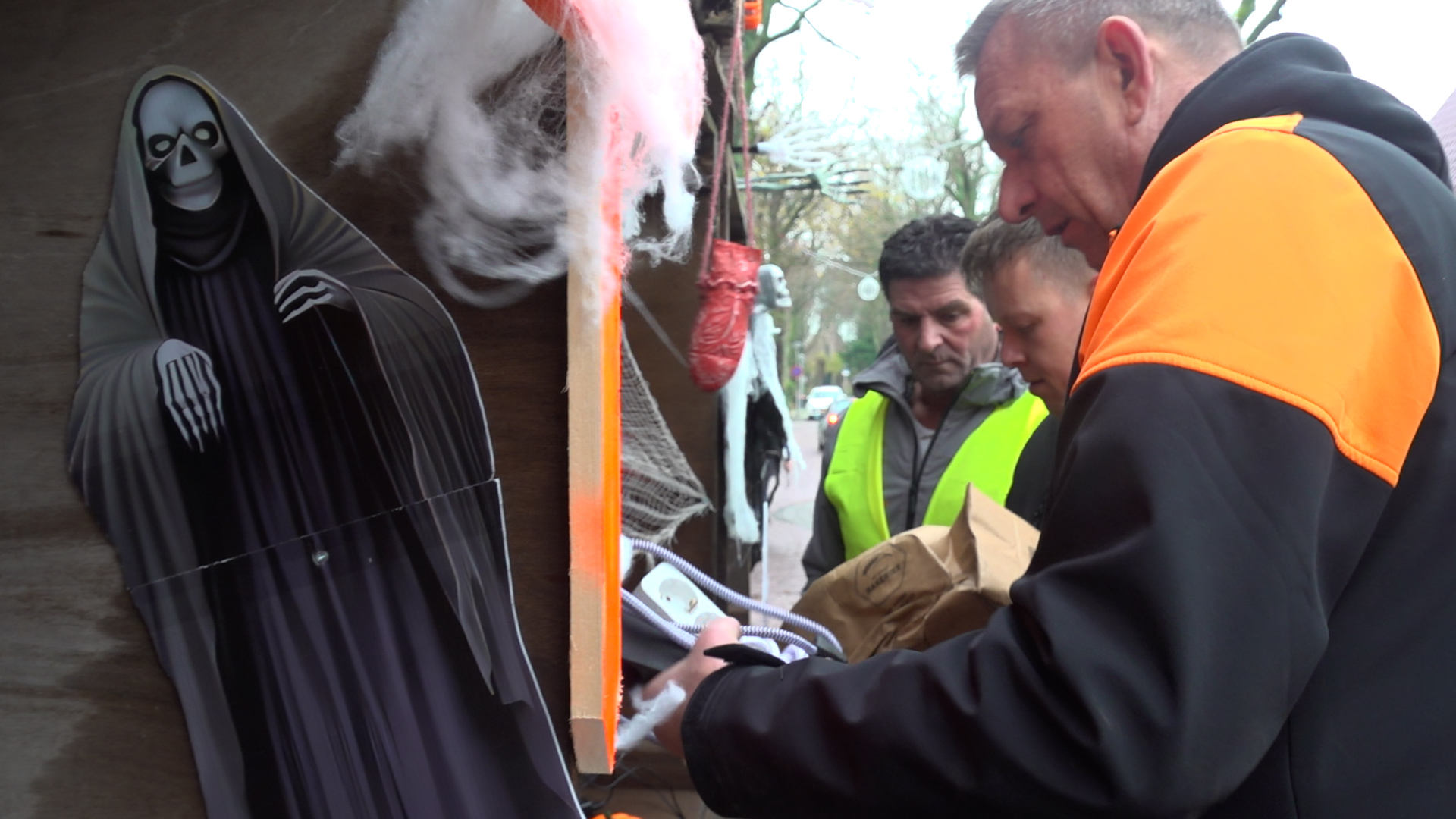 Drie mannen in oranje en gele veiligheidsjassen inspecteren Halloween-decoraties, waaronder een skeletfiguur en spinnenwebben.