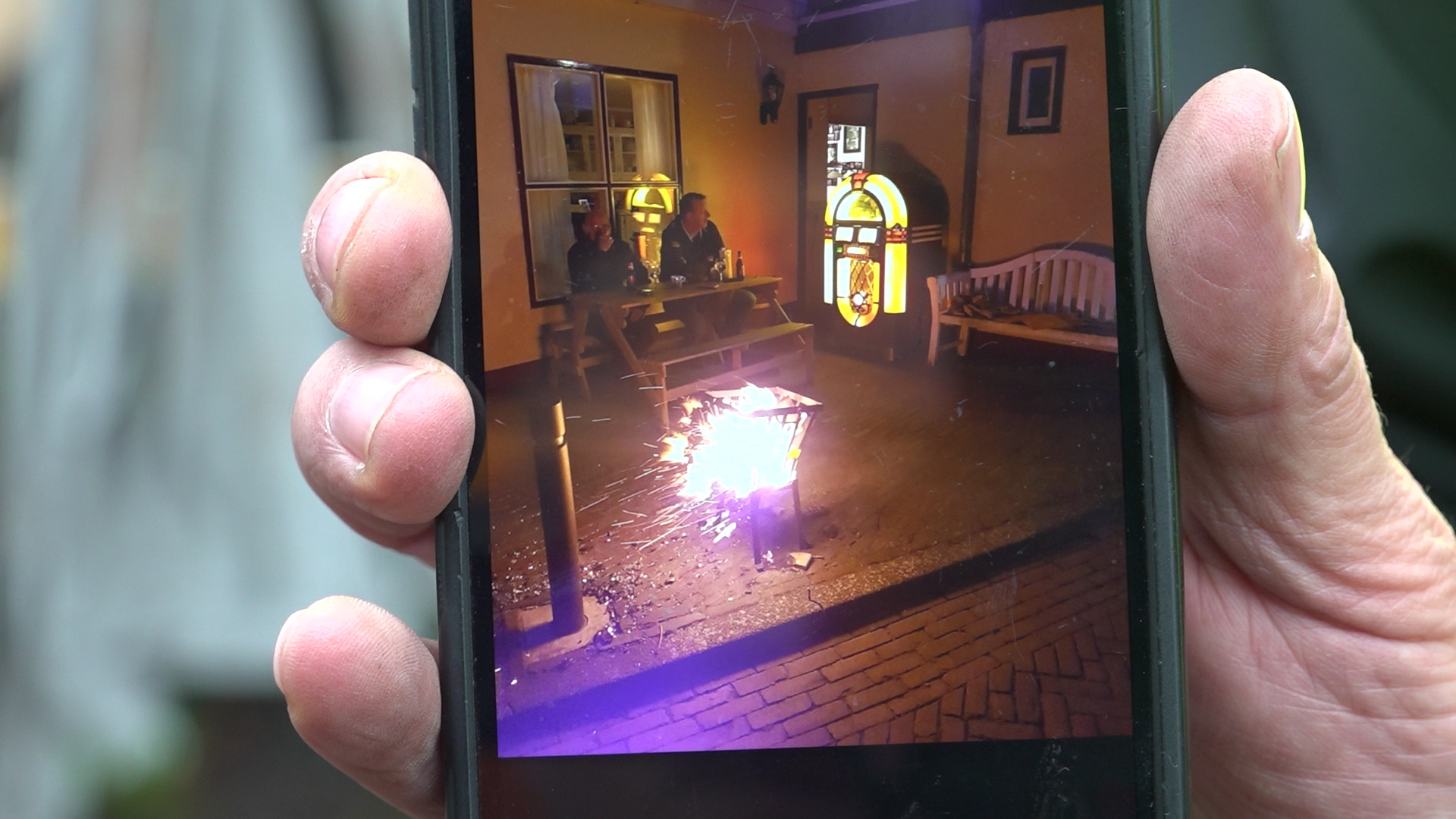 Een hand houdt een smartphone vast met daarop een afbeelding van een veranda, waar een smeulend vuur in een vuurkorf en een fel verlichte jukebox te zien zijn met twee mensen die aan een tafel zitten in een gezellige sfeer.