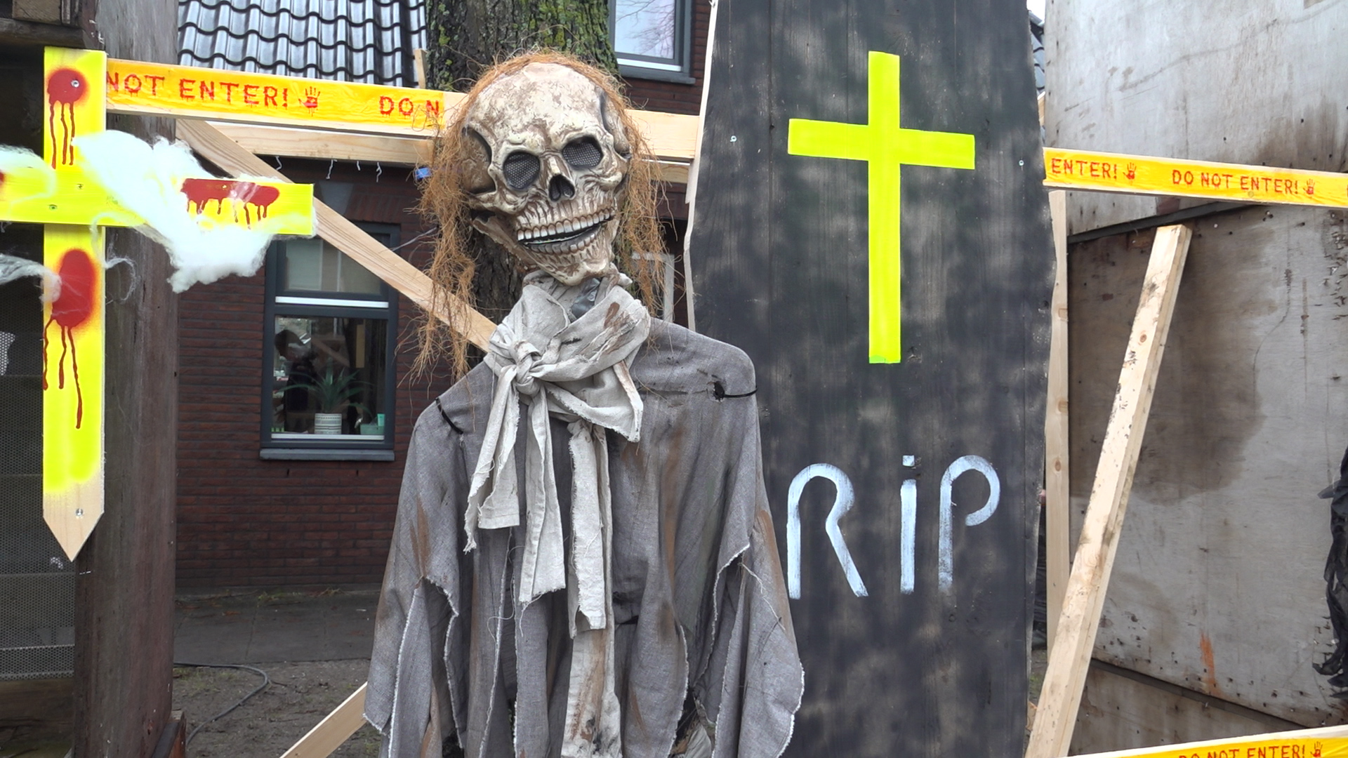Halloween-decoratie met een skeletfiguur in oude kleding en een groot kruisbord met RIP erop geschilderd, omringd door gele linten met 'Do Not Enter!' waarschuwingen.