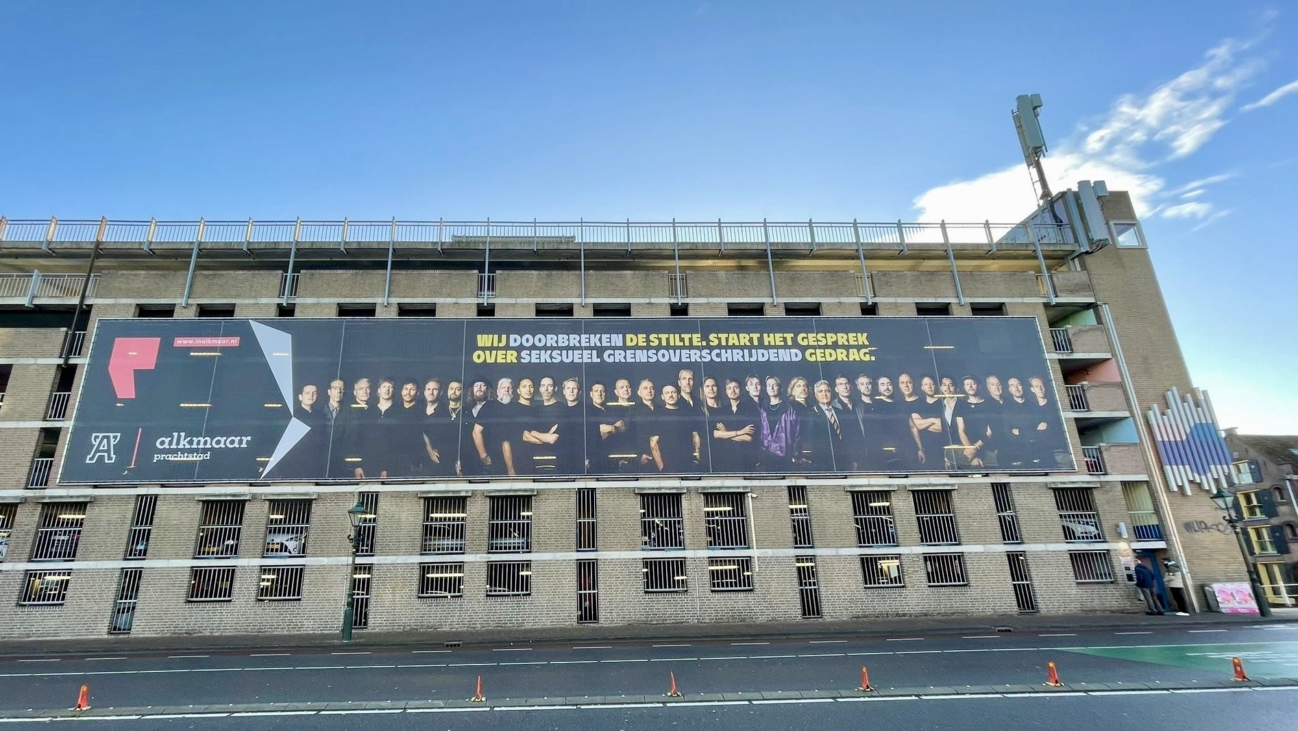 Een groot billboard op de zijkant van een gebouw toont een groep mensen in zwarte kleding met een tekst in het Nederlands, "Wij doorbreken de stilte. Start het gesprek over seksueel grensoverschrijdend gedrag." Het gebouw en de heldere lucht zijn zichtbaar.