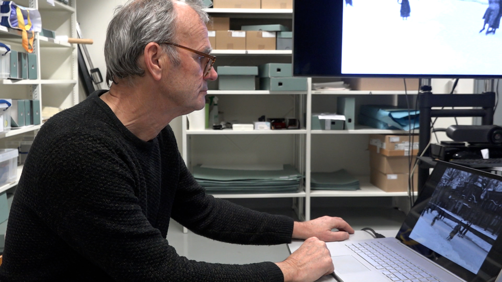Een man met bril zit aan een bureau en kijkt naar een laptop waarop een afbeelding van een besneeuwd landschap te zien is; op de achtergrond staan planken met dozen in een archiefruimte.