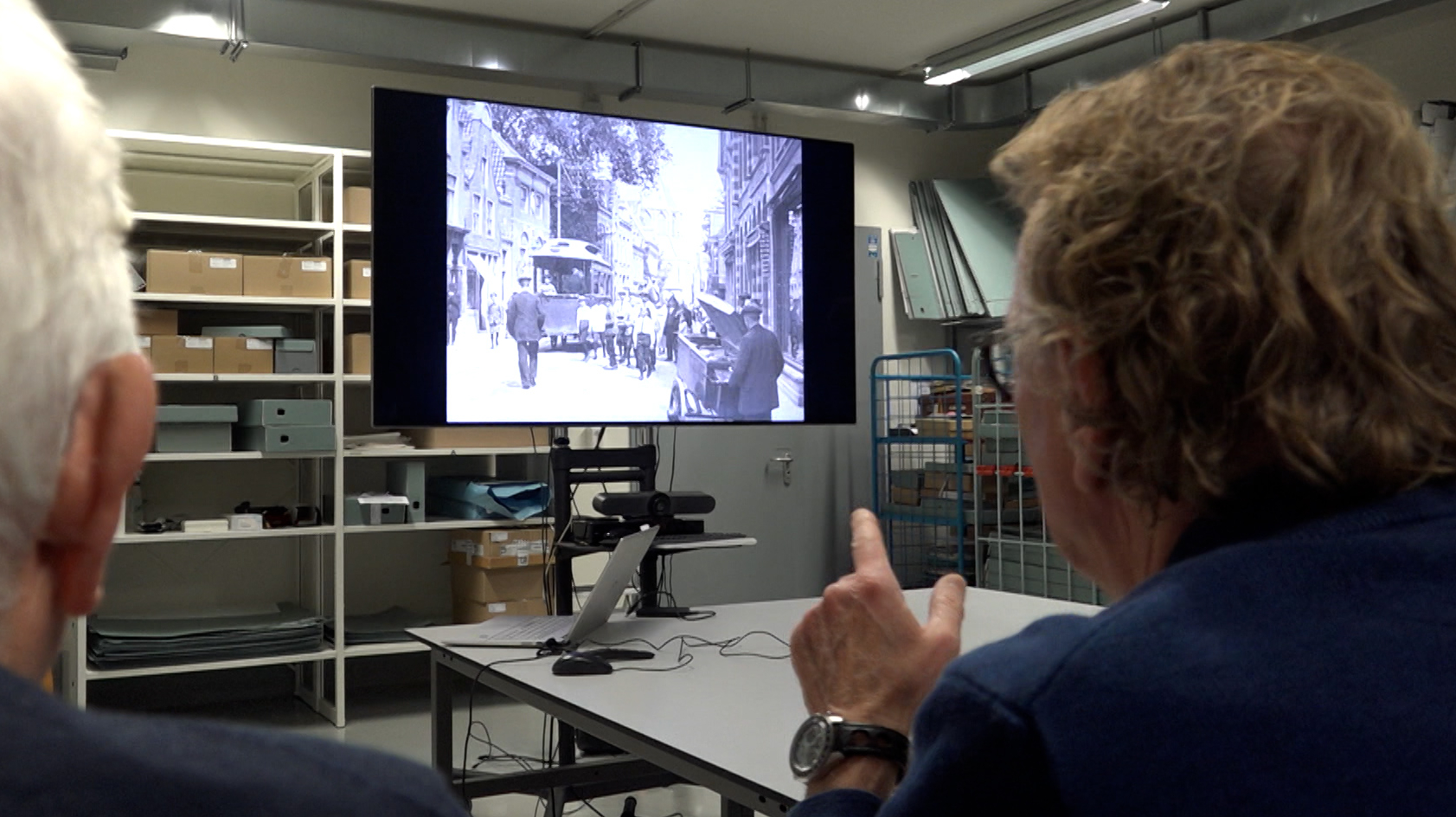Twee mensen kijken naar een zwart-witfoto van een historische stadsstraat, afgebeeld op een scherm in een archiefruimte met planken vol dozen en documenten.