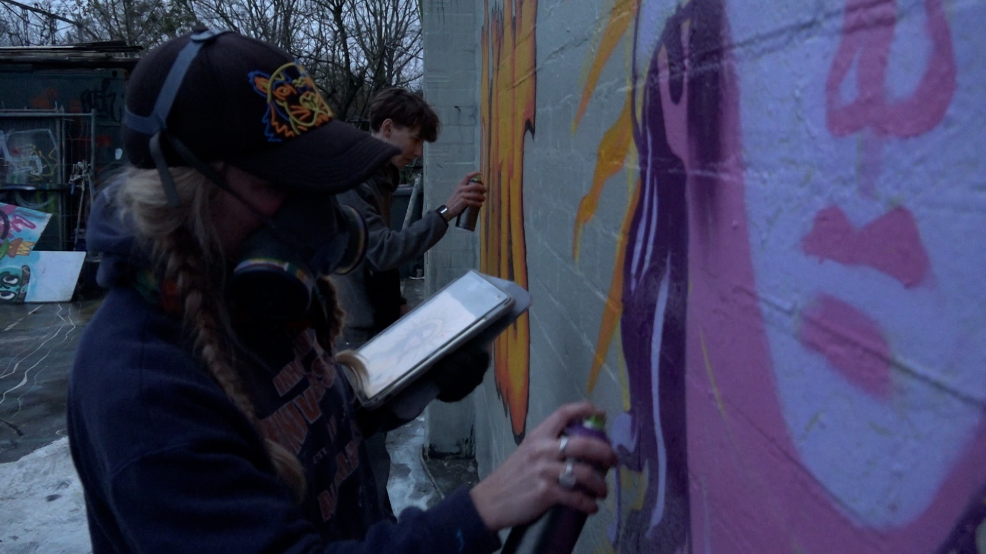 Twee personen zijn buiten een muur aan het verven met spuitbussen; de voorste persoon draagt een pet en een gasmasker, terwijl de ander op de achtergrond werkt.